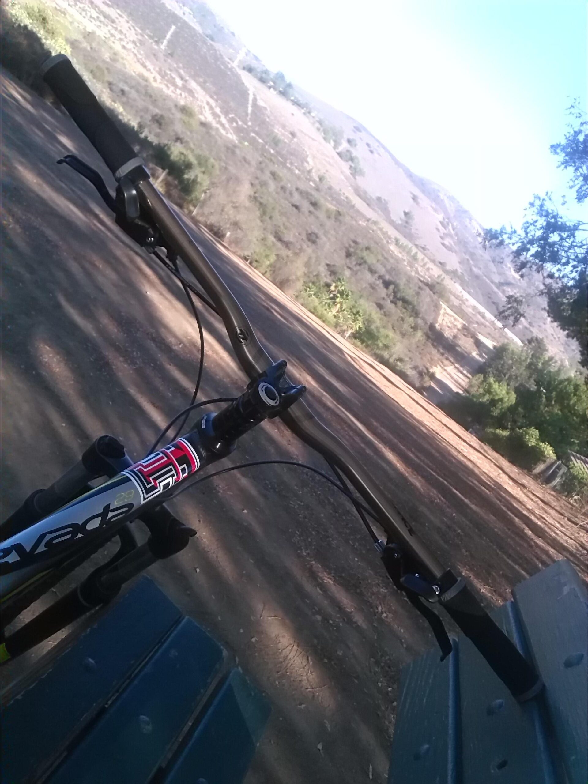 Fuji Nevada 29 1.0: Close-up view of a mountain bike's handlebars, with a wooded trail and hills in the background. The bike is resting on a green bench, indicating a moment of pause amidst a ride in nature.