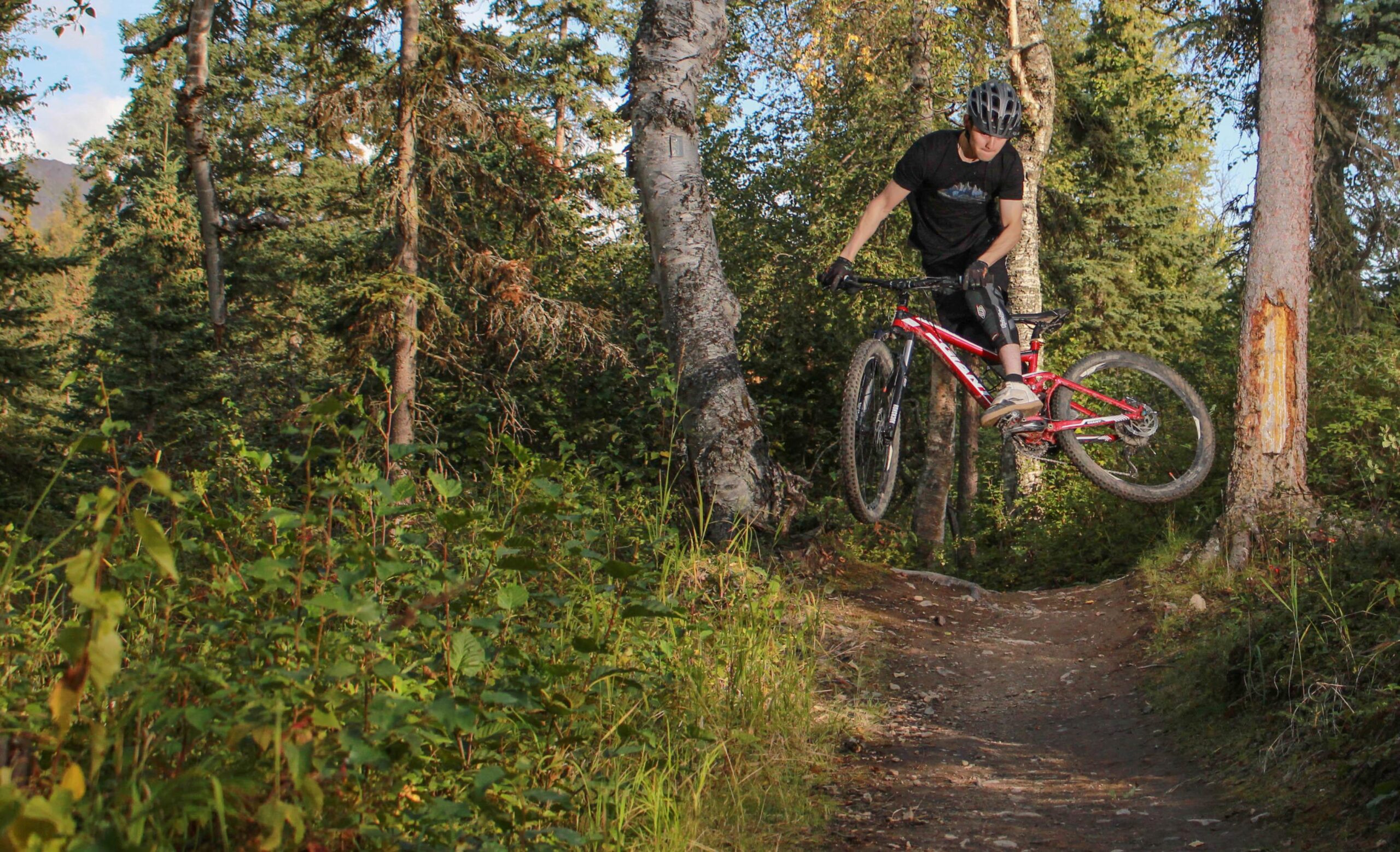 A mountain biker performing a jump on a dirt trail surrounded by tall trees and greenery. The cyclist is wearing a helmet and biking gear, showcasing mid-air action with the bike raised off the ground. Sta Trails mountain bike trail.