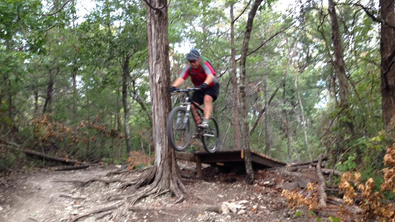 A mountain biker in a red shirt and helmet jumps off a wooden ramp in a forested area, surrounded by trees and rocky terrain. Boulder Park mountain bike trail.