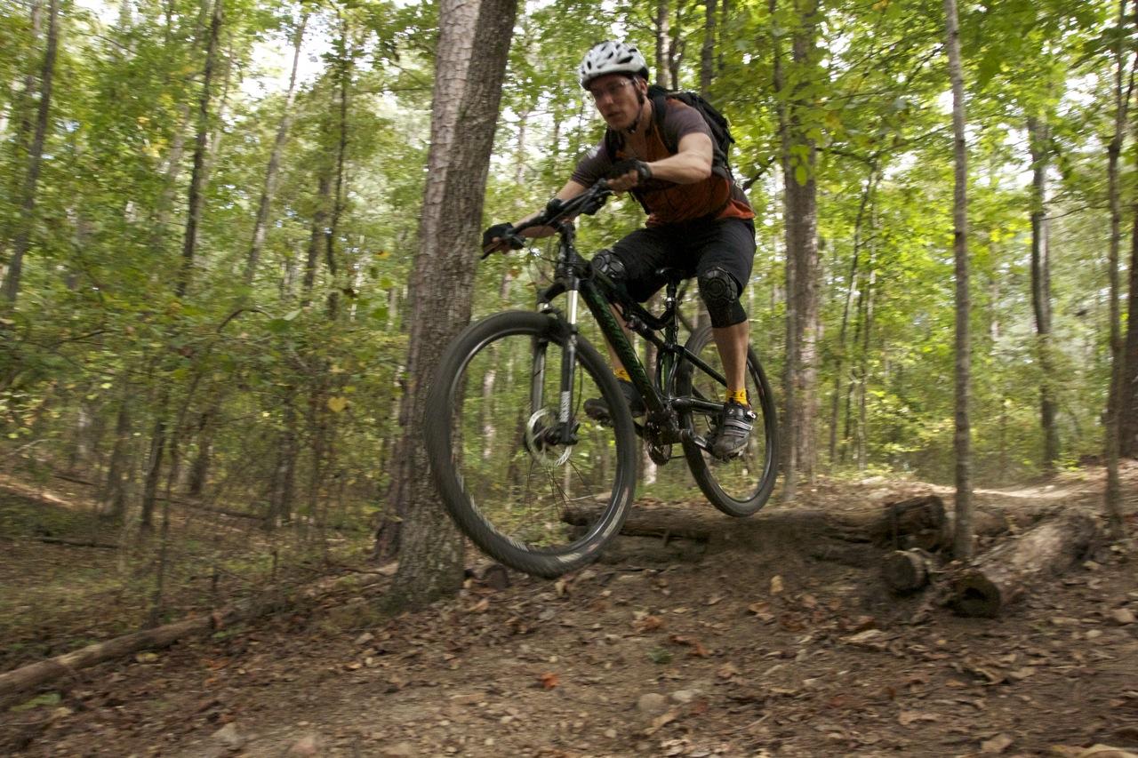 A mountain biker jumping over a small log on a wooded trail, surrounded by trees and greenery. The cyclist is wearing a helmet and protective gear, showcasing an action-packed moment in outdoor sports. Blankets Creek mountain bike trail.