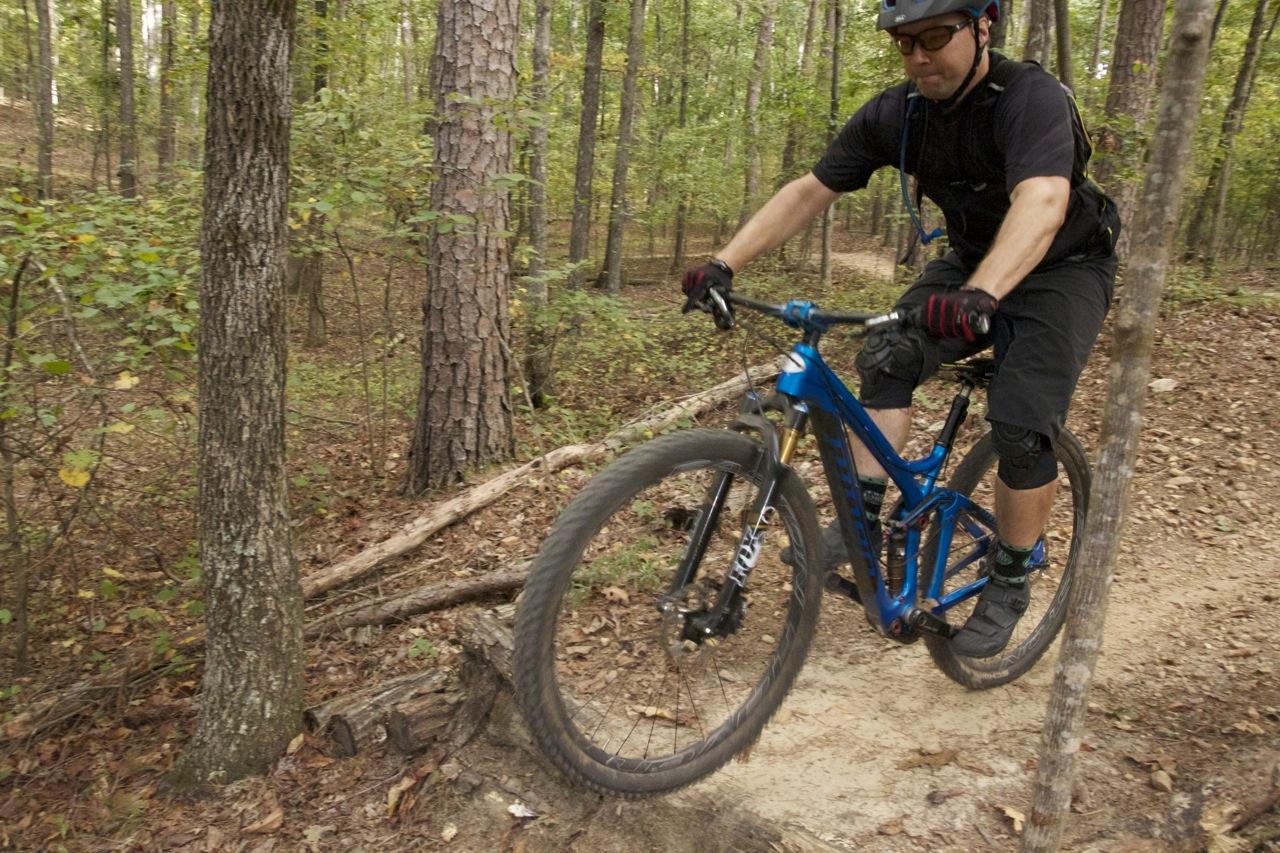 A mountain biker riding through a forest trail, navigating over a small log with one wheel off the ground, wearing a helmet and protective gear. The surrounding landscape features tall trees and earthy foliage typical of a wooded area. Blankets Creek mountain bike trail.