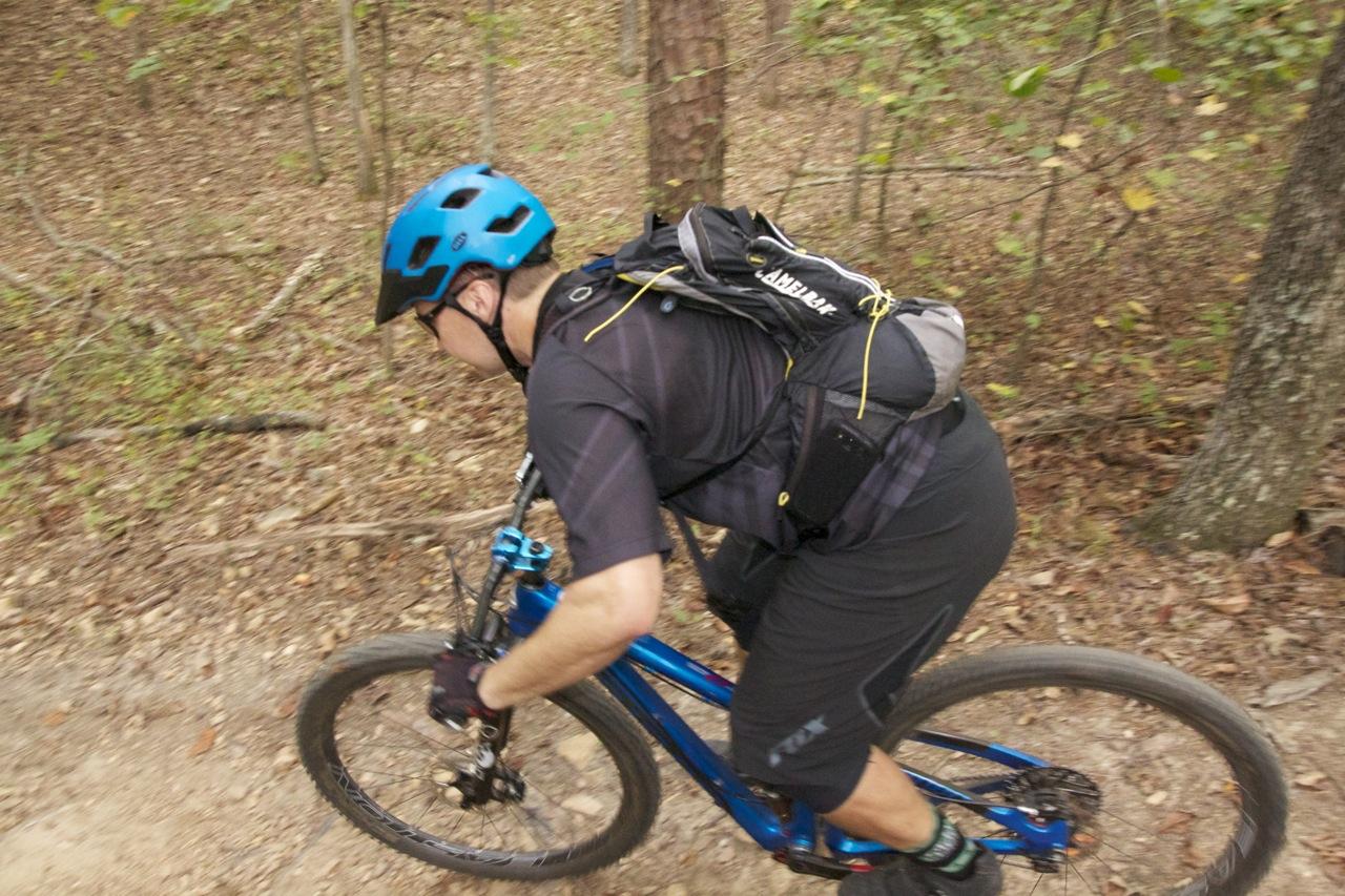 A cyclist wearing a blue helmet and a black backpack rides a blue mountain bike along a dirt trail in a wooded area. The cyclist leans forward as they navigate the uneven terrain, surrounded by trees and foliage. Blankets Creek mountain bike trail.