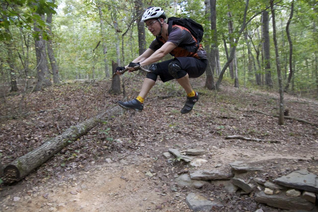 A mountain biker wearing a helmet and protective gear performs a jump over a fallen log on a forest trail, surrounded by trees and foliage. Blankets Creek mountain bike trail.