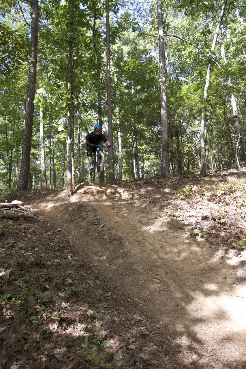 A mountain biker performing a jump on a dirt trail surrounded by trees in a wooded area. The rider is wearing a helmet and riding gear, capturing a moment of action and adventure in nature. Blankets Creek mountain bike trail.