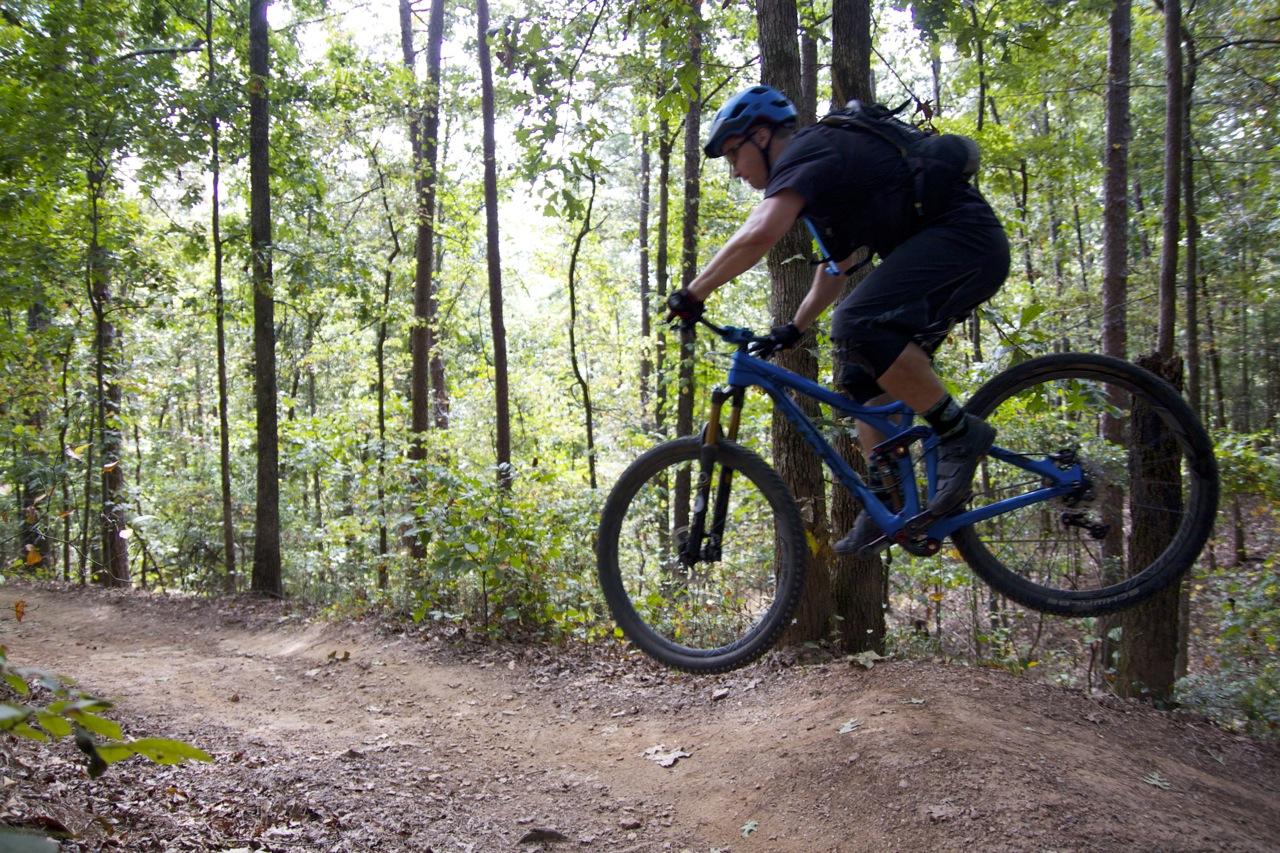 A mountain biker in a blue helmet and black attire jumps off a dirt ramp surrounded by trees and greenery. The rider is captured mid-air, demonstrating a dynamic action pose as they navigate a wooded trail. Blankets Creek mountain bike trail.