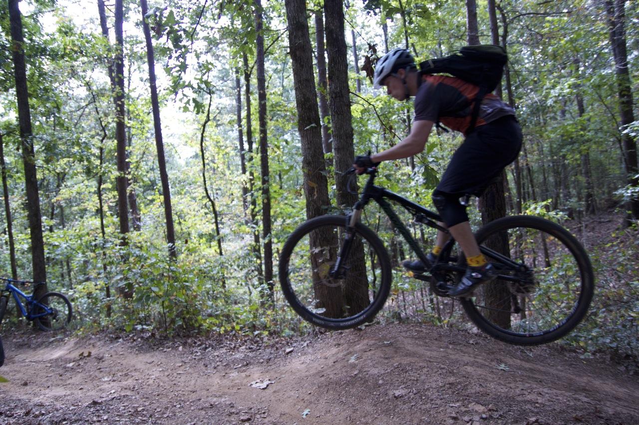 A mountain biker in mid-air jumps off a dirt ramp on a forest trail, surrounded by tall trees and dense greenery. Blankets Creek mountain bike trail.