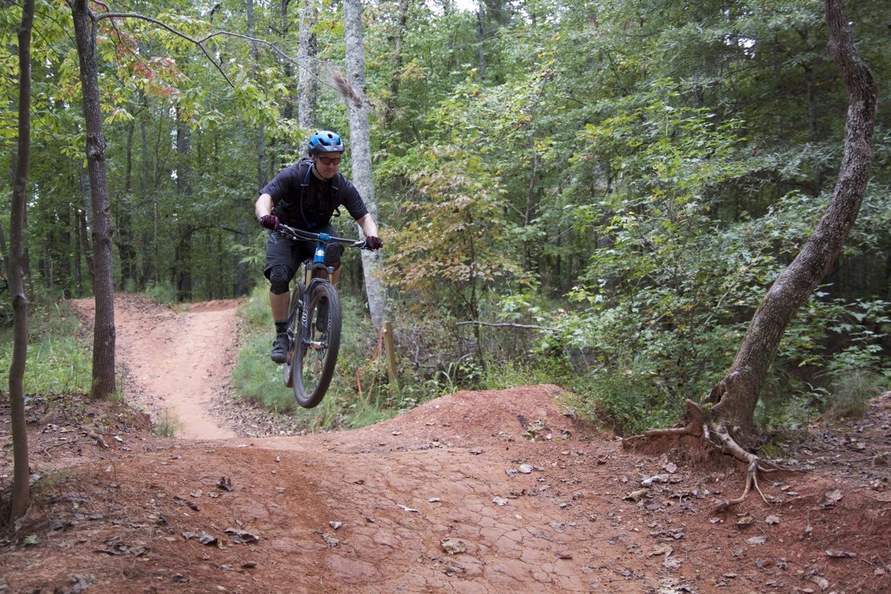 A person riding a mountain bike jumps over a dirt ramp on a forest trail. The cyclist is wearing a blue helmet and protective gear, with trees and a natural landscape visible in the background. Blankets Creek mountain bike trail.