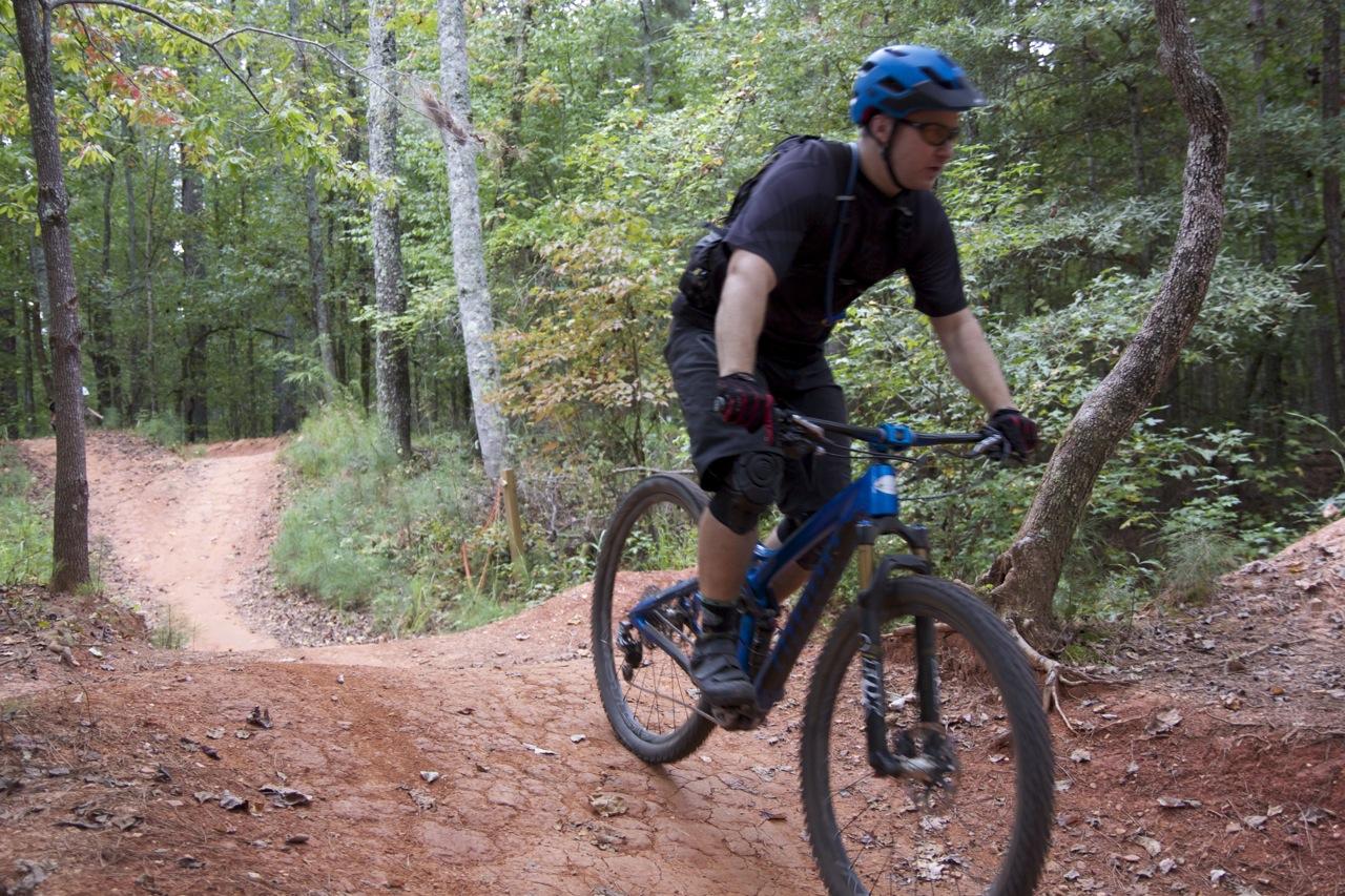 A mountain biker wearing a blue helmet and protective gear rides along a dirt trail in a wooded area, navigating a bend. The surrounding trees are lush and green, indicating a natural setting for outdoor biking. Blankets Creek mountain bike trail.