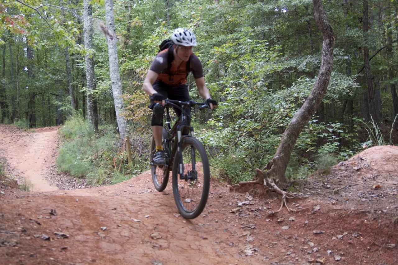 A cyclist navigating a dirt mountain bike trail in a wooded area, wearing a helmet and orange jersey, with trees and greenery surrounding the path. Blankets Creek mountain bike trail.
