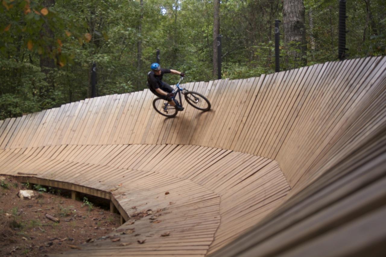 A person riding a mountain bike on a wooden pump track, leaning into a turn surrounded by trees in a natural forest setting. The track features a series of curved wooden ramps, highlighting the dynamics of off-road cycling. Blankets Creek mountain bike trail.