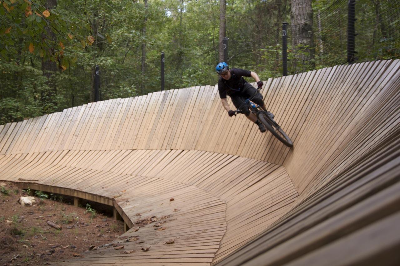 A mountain biker navigating a wooden pump track in a forest, leaning into a curve while riding on a smoothly constructed wooden surface surrounded by trees. Blankets Creek mountain bike trail.