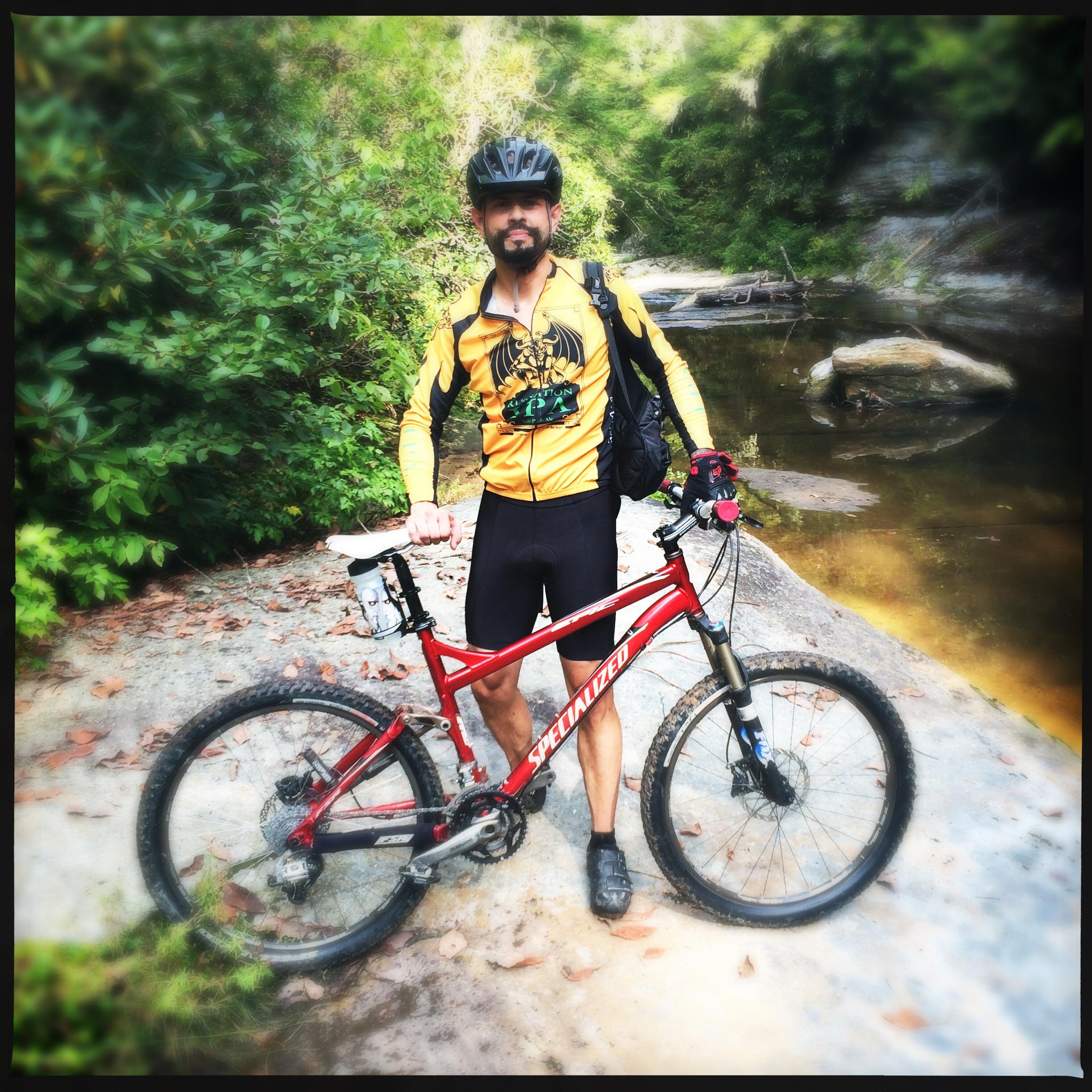 A mountain biker wearing a yellow jersey and black shorts stands beside a red Specialized mountain bike on a rocky riverbank, surrounded by lush greenery. A small stream is visible in the background, reflecting the natural environment. Sheltowee Trace - Laurel Lake Trail mountain bike trail.