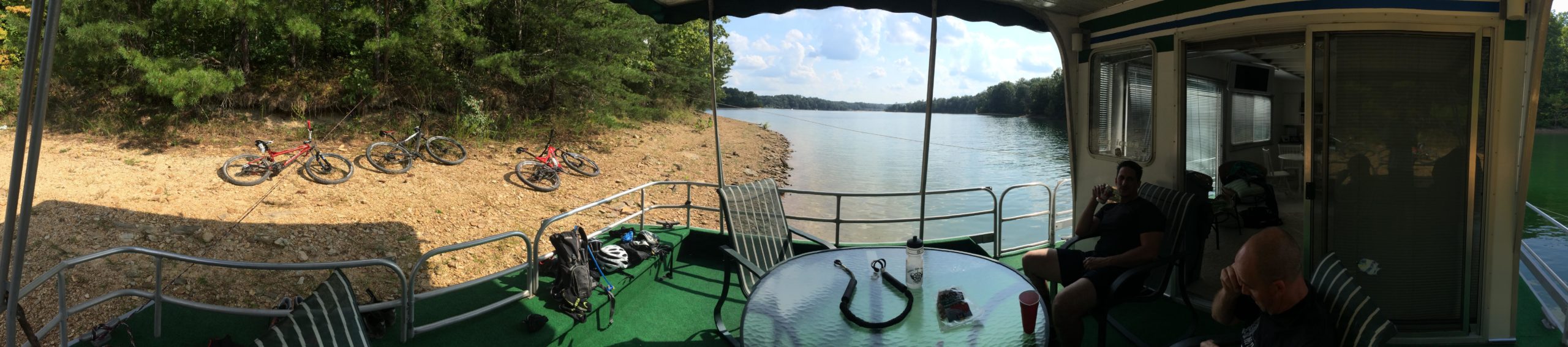A panoramic view from inside a houseboat, featuring a green outdoor area with a table and chairs. In the background, several bicycles are parked on the shore of a calm lake surrounded by trees. There are two people sitting at the table, enjoying the scenic view under a partly cloudy sky. Sheltowee Trace - Laurel Lake Trail mountain bike trail.