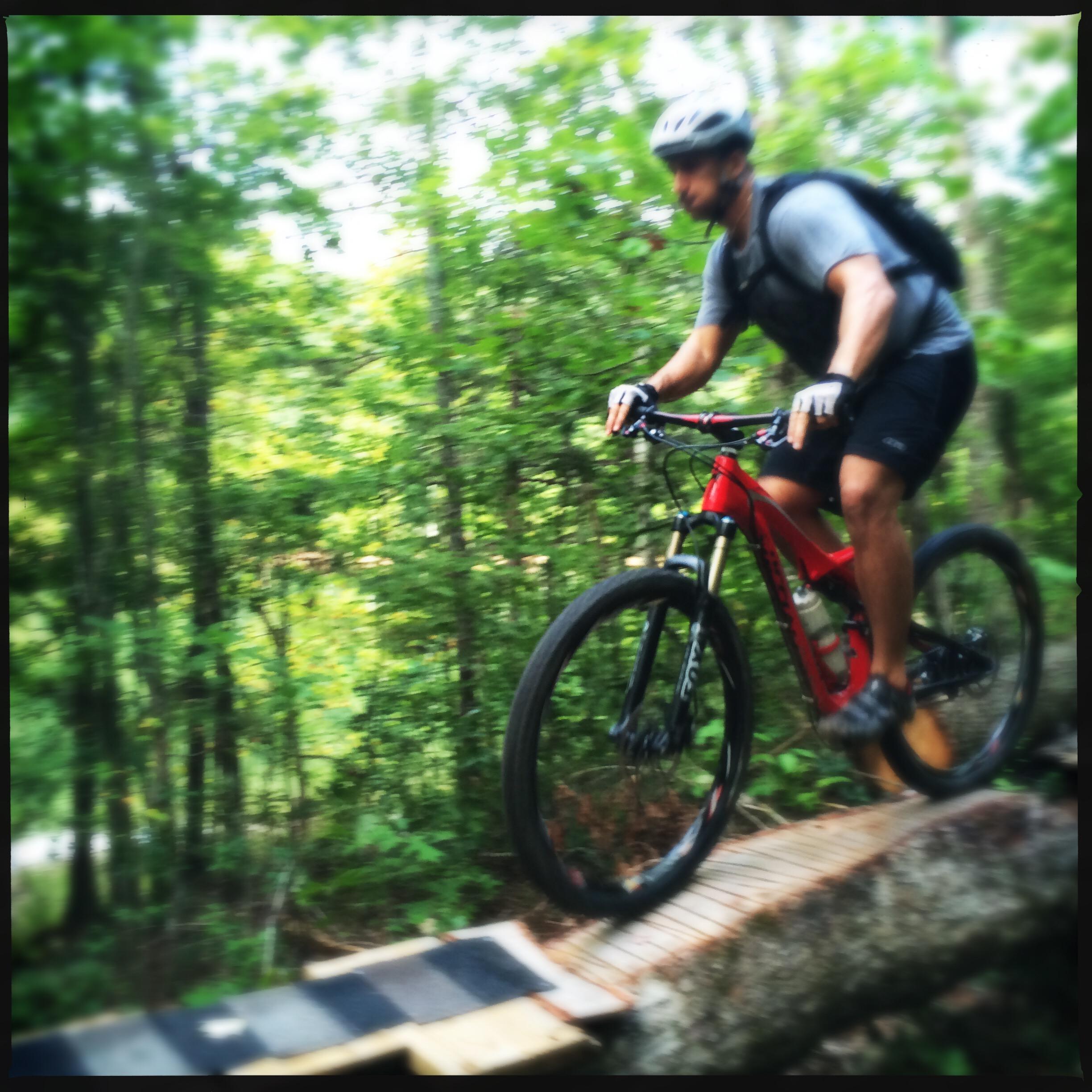 A mountain biker navigating a wooden bridge over rough terrain in a dense forest. The rider is wearing a helmet and a backpack, showcasing a dynamic posture as they pedal along the trail. The scene captures the adventure and thrill of outdoor cycling. Sheltowee Trace - Laurel Lake Trail mountain bike trail.