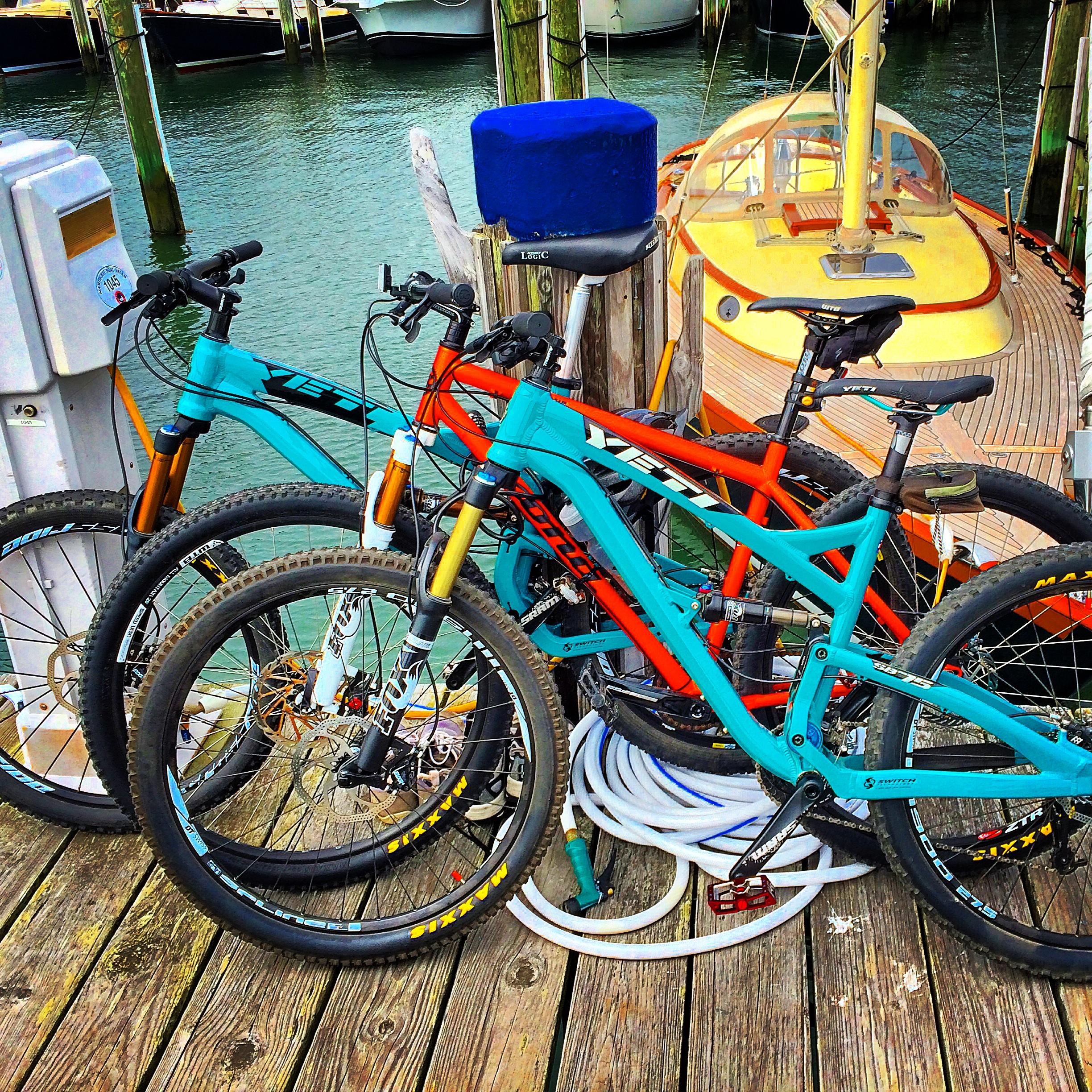 Yeti SB-75: Two mountain bikes, one turquoise and one orange, are leaning against a wooden dock with a blue cushion nearby. In the background, a small boat is moored on calm water, and there are ropes and a hose on the dock. The scene reflects a lively harbor atmosphere.