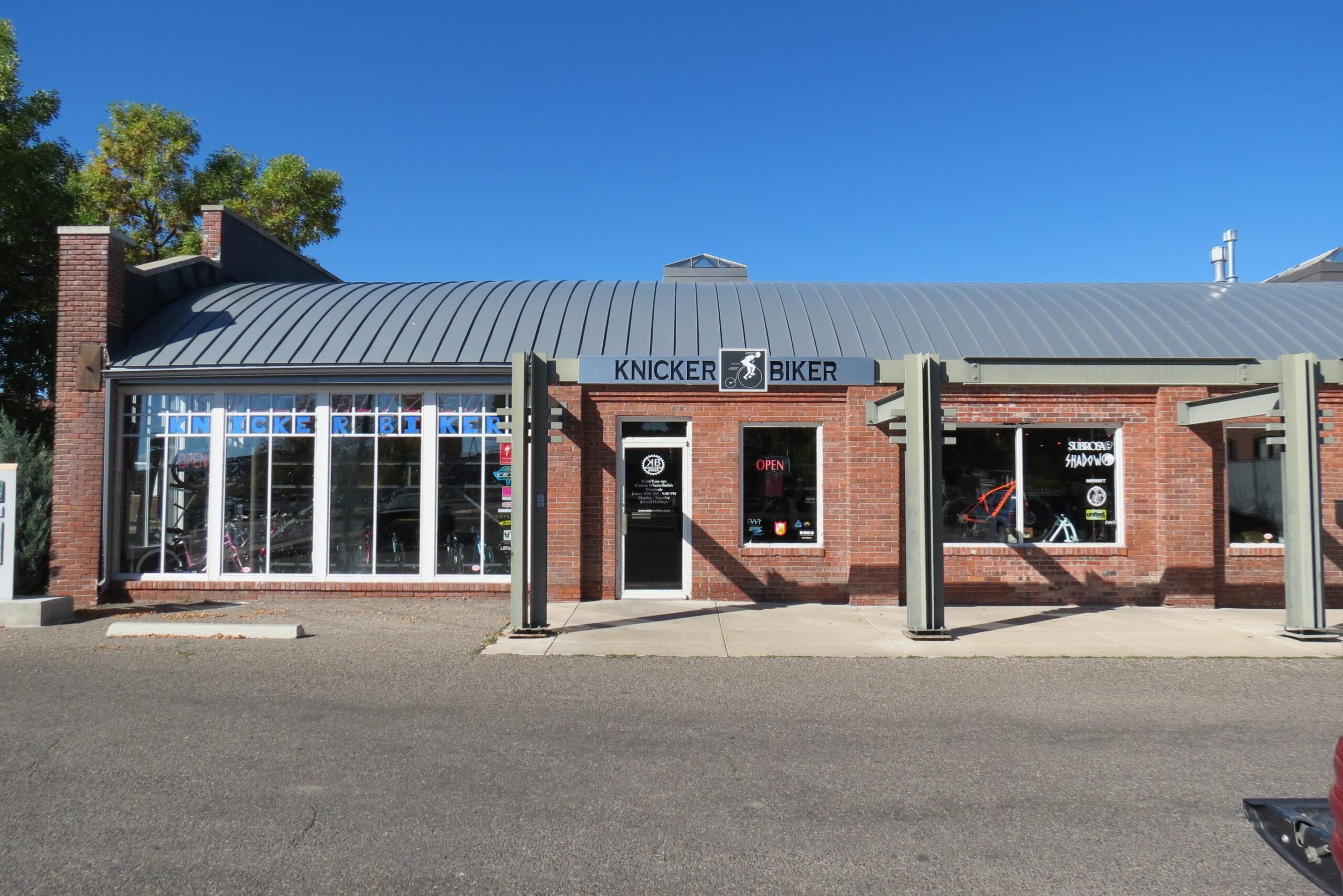 A brick storefront featuring a sign that reads "KNICKER BIKER." The building has large windows displaying various biking gear and bicycles. The entrance has a door with an "OPEN" sign visible. It has a metal roof and is surrounded by a parking area and some greenery. The sky is clear and blue, indicating a sunny day.