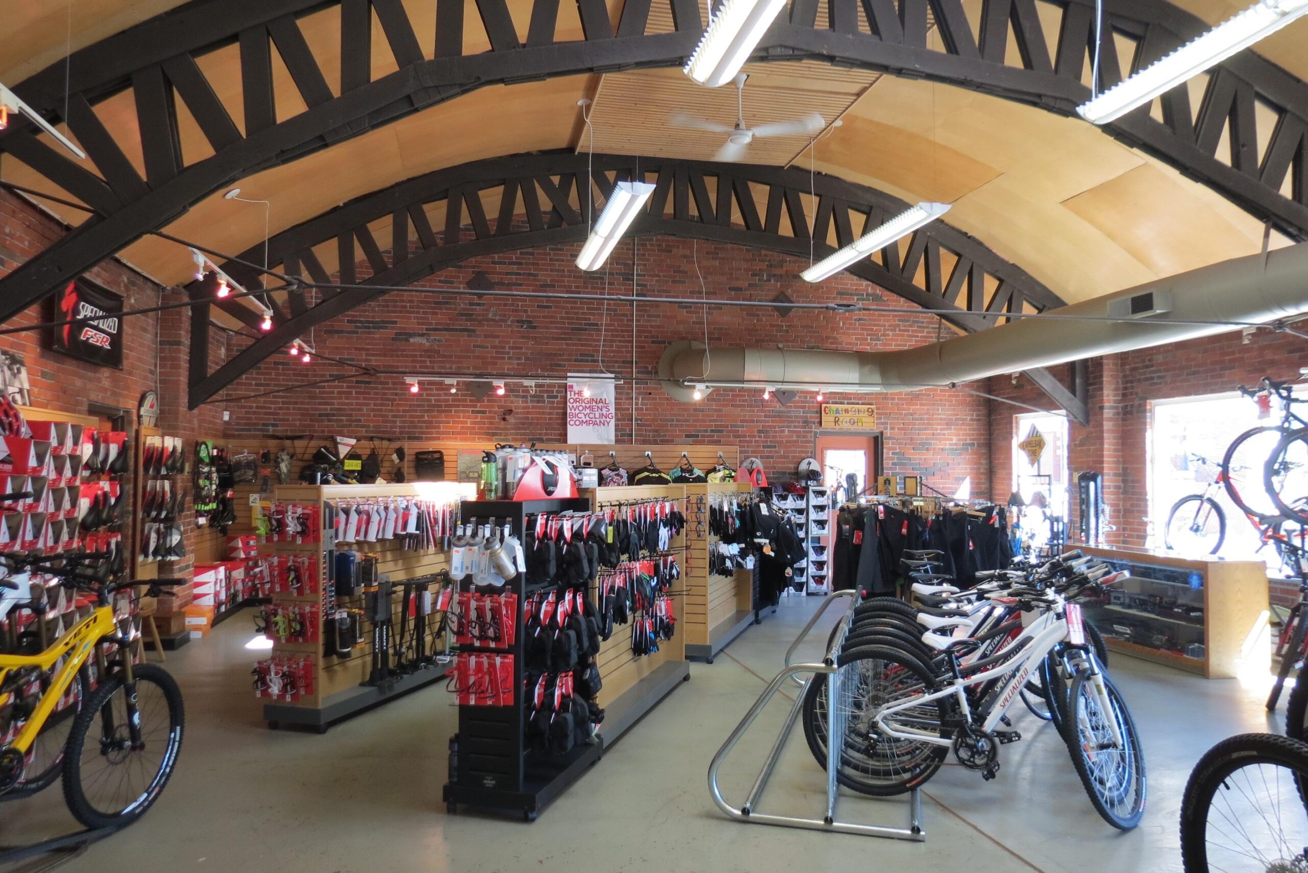 Interior of a bike shop featuring high arched wooden ceilings, exposed brick walls, and various bicycle accessories displayed on wooden shelving units. Several mountain bikes and road bikes are lined up for sale, alongside racks of cycling gear, helmets, and other biking equipment. Bright overhead lights and a large window provide ample illumination.