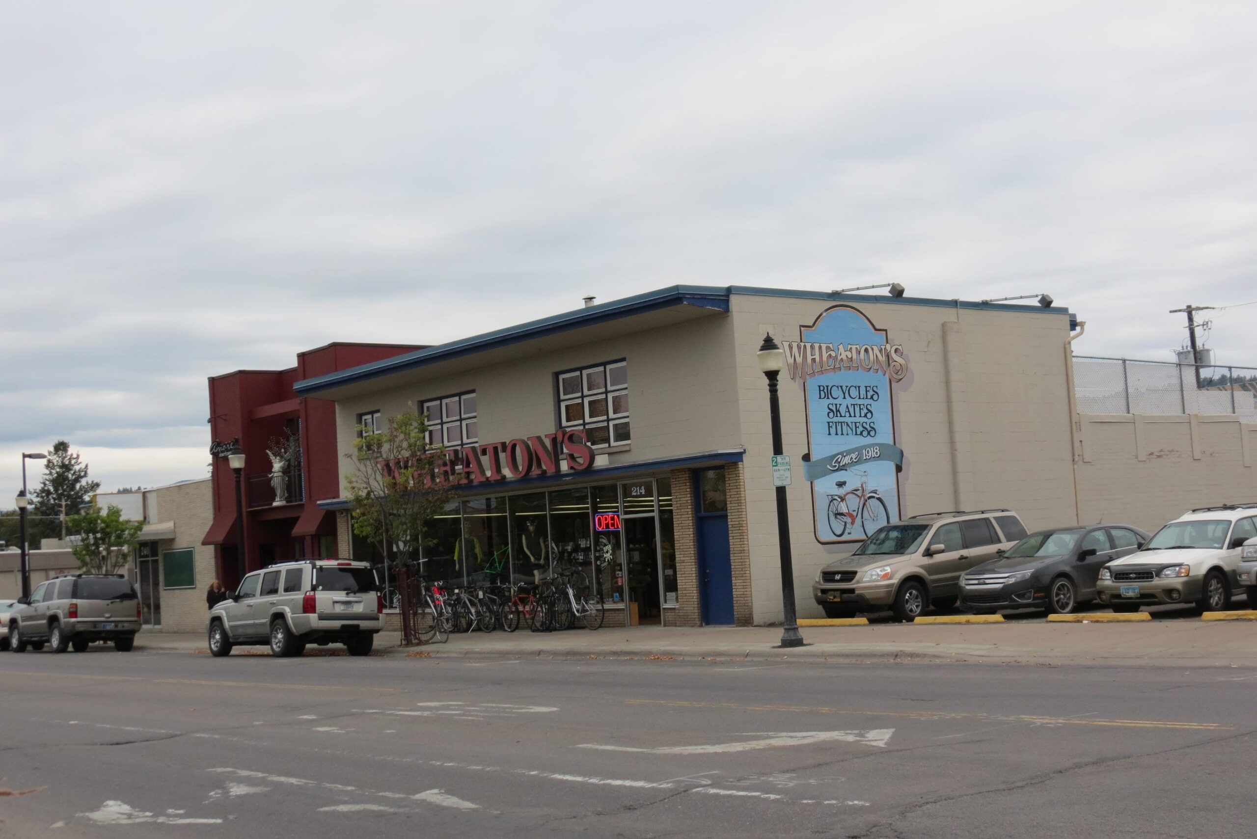 Image of a storefront for "Wheaton's," a shop specializing in bicycles, skates, and fitness equipment. The building features large windows displaying bicycles and an "Open" sign. A blue and white sign highlights the store's offerings and its establishment since 1913. Parked vehicles line the street in front of the store, and cloudy skies are visible above.