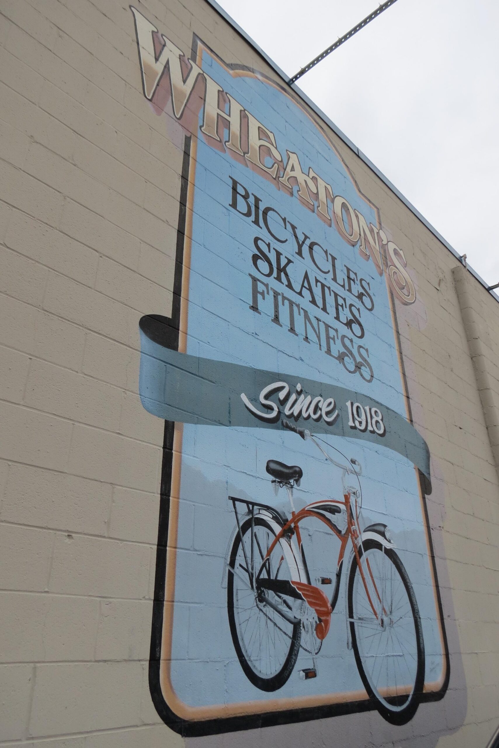 Mural featuring a vintage-style bicycle with the text "Wheaton's Bicycles Skates Fitness Since 1918" in decorative lettering, painted on a brick wall.