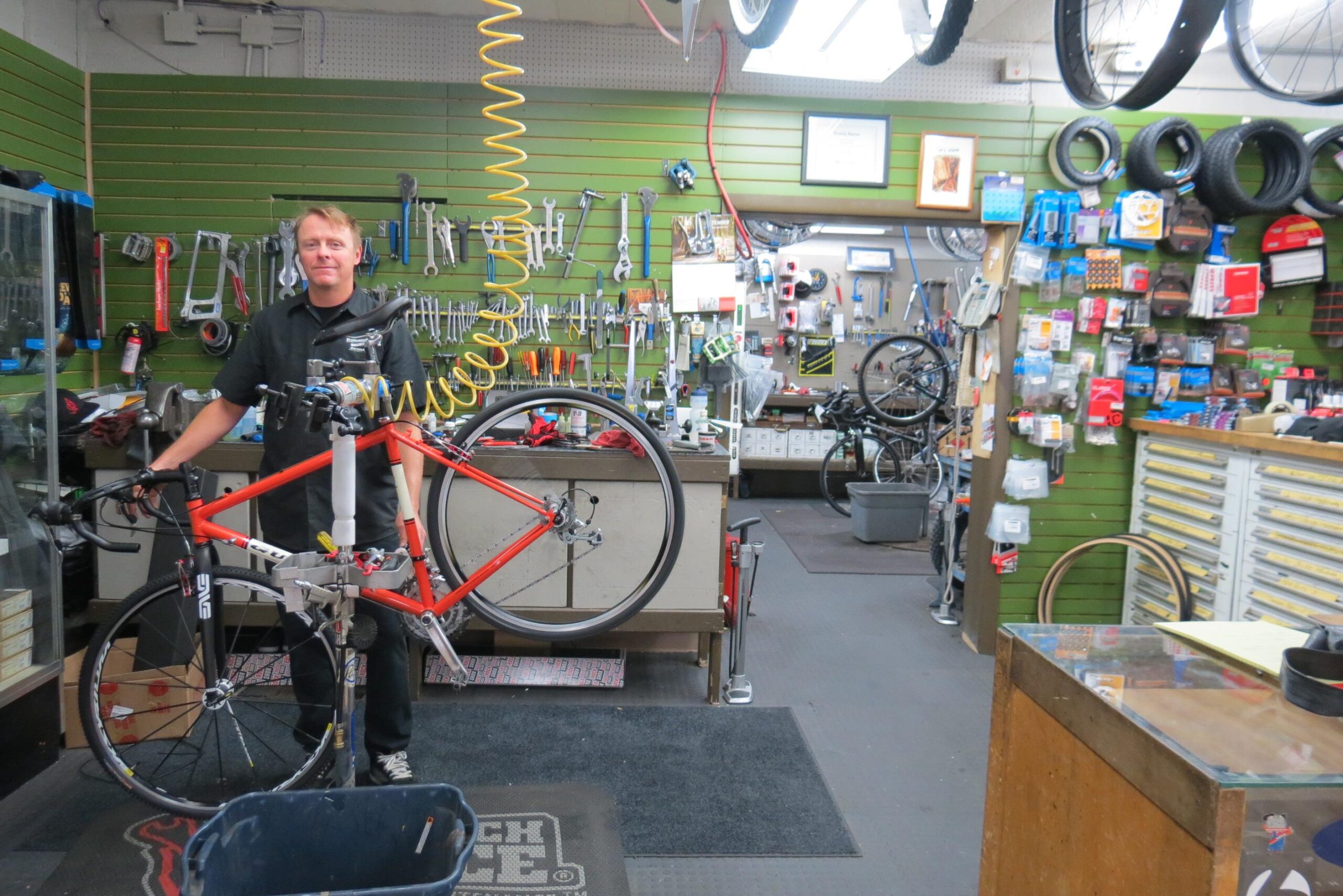 A bike mechanic stands in a well-organized workshop, holding an orange bicycle that is mounted on a repair stand. The background features various tools, bike parts, and accessories displayed on green walls, along with several bicycles in the shop. The floor is covered with a dark mat, and a table in the foreground holds additional equipment.