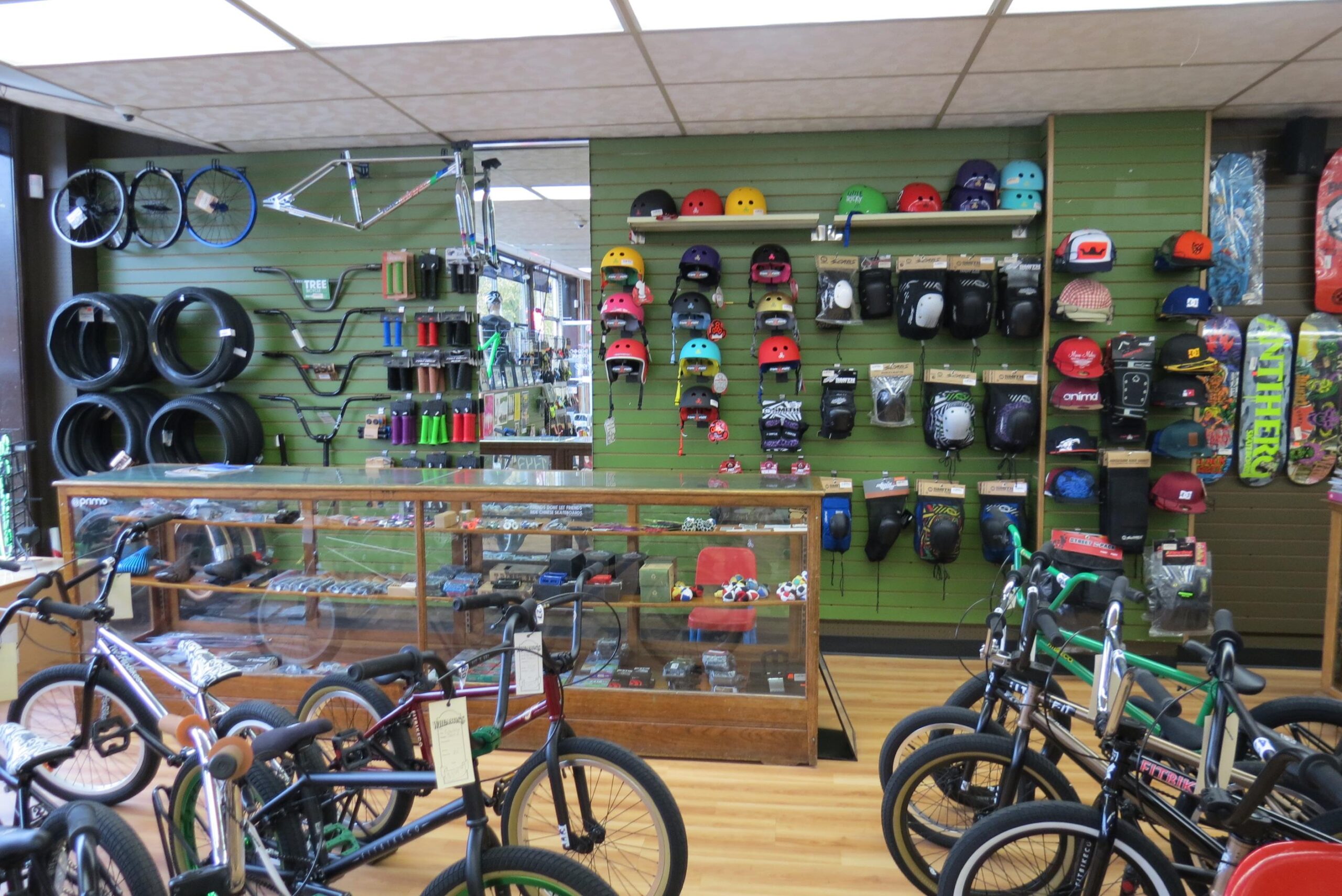 A bicycle shop interior featuring a display of BMX bikes in the foreground, various helmets, pads, and accessories hanging on the green wall behind. Shelves showcase bicycle parts like tires and handlebars, as well as a glass display case containing additional equipment. The shop has a bright, inviting atmosphere with wooden flooring.