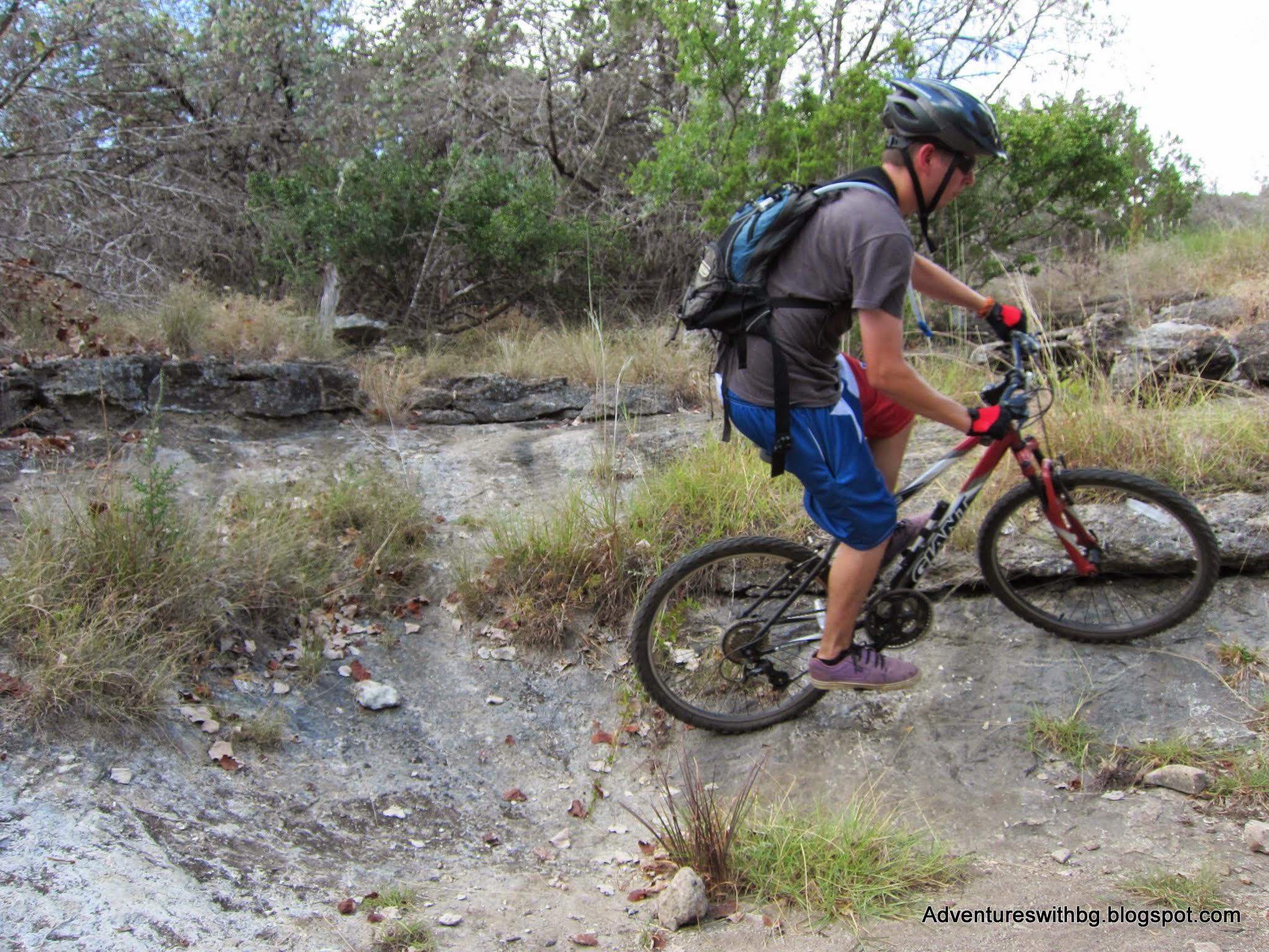 A person riding a mountain bike on a rocky trail surrounded by dry grass and sparse trees. The cyclist is wearing a helmet, gloves, and a backpack, navigating a steep incline. Goodwater Trail mountain bike trail.