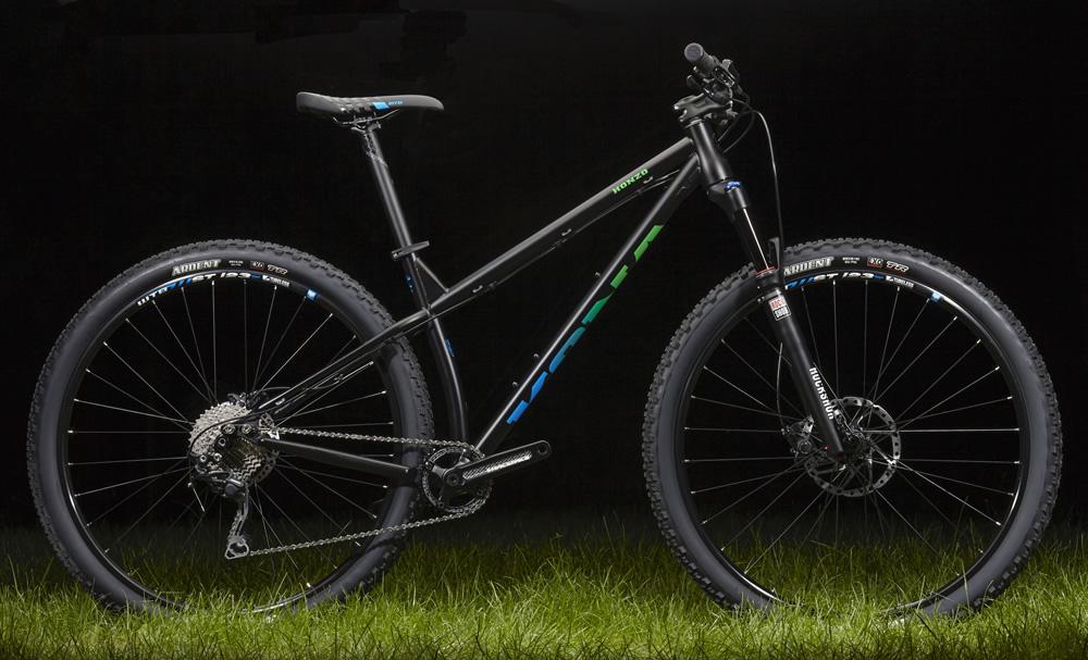 Kona Honzo: A black mountain bike with blue and green accents displayed on grass, showcasing detailed components including disc brakes and knobby tires, against a dark background.