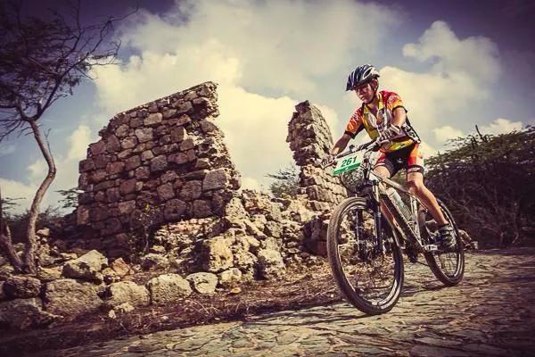 Trek 8000: A cyclist riding a mountain bike on a cobblestone path, passing by ancient stone ruins with a backdrop of a partly cloudy sky. The rider is wearing a colorful jersey and a helmet, showcasing an active outdoor scene.