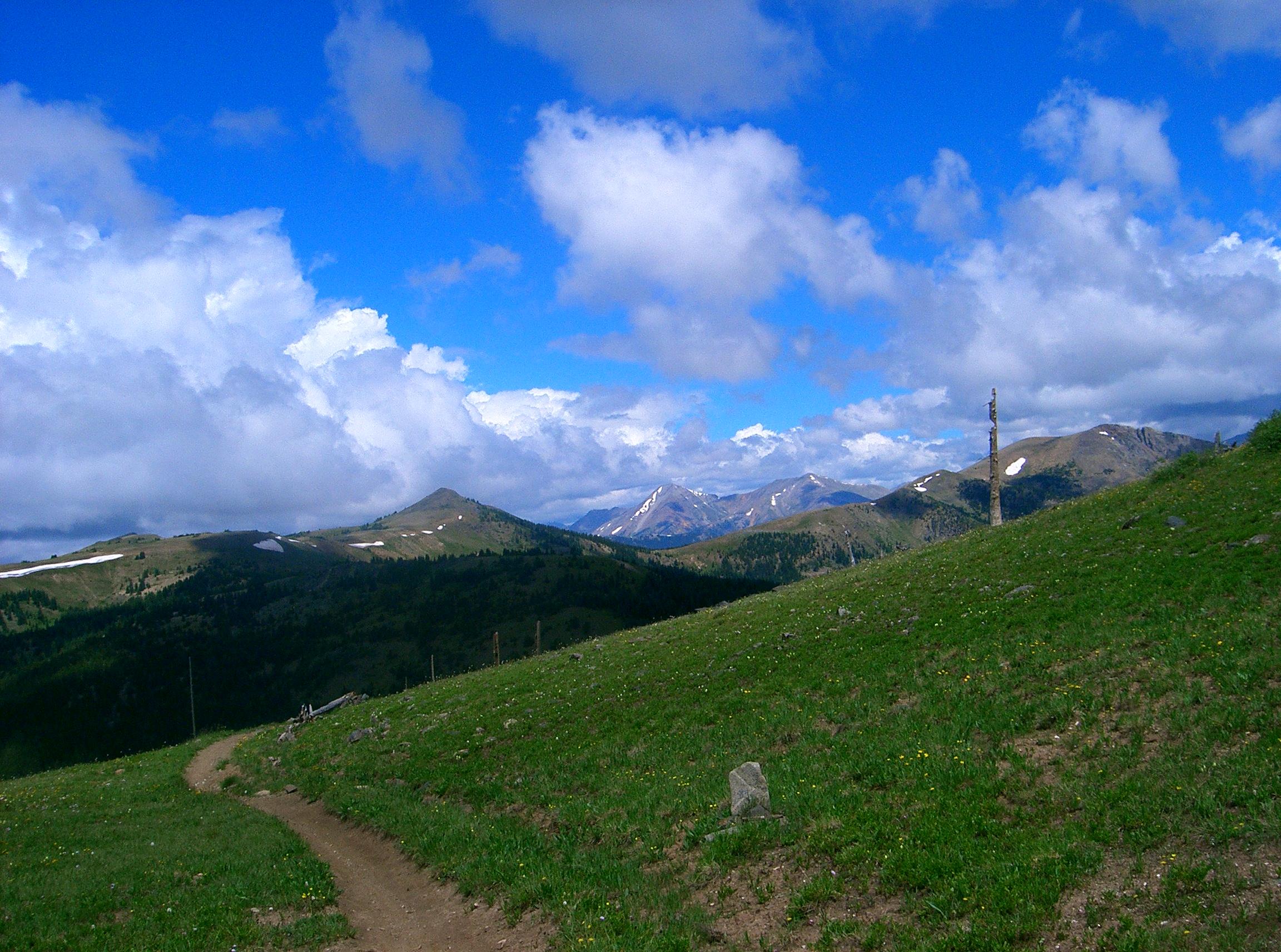 A winding dirt path leads through a lush green landscape, surrounded by rolling hills and majestic mountains under a partly cloudy blue sky. Snow-capped peaks are visible in the distance, adding to the scenic beauty of the natural setting. Monarch Crest Trail mountain bike trail.