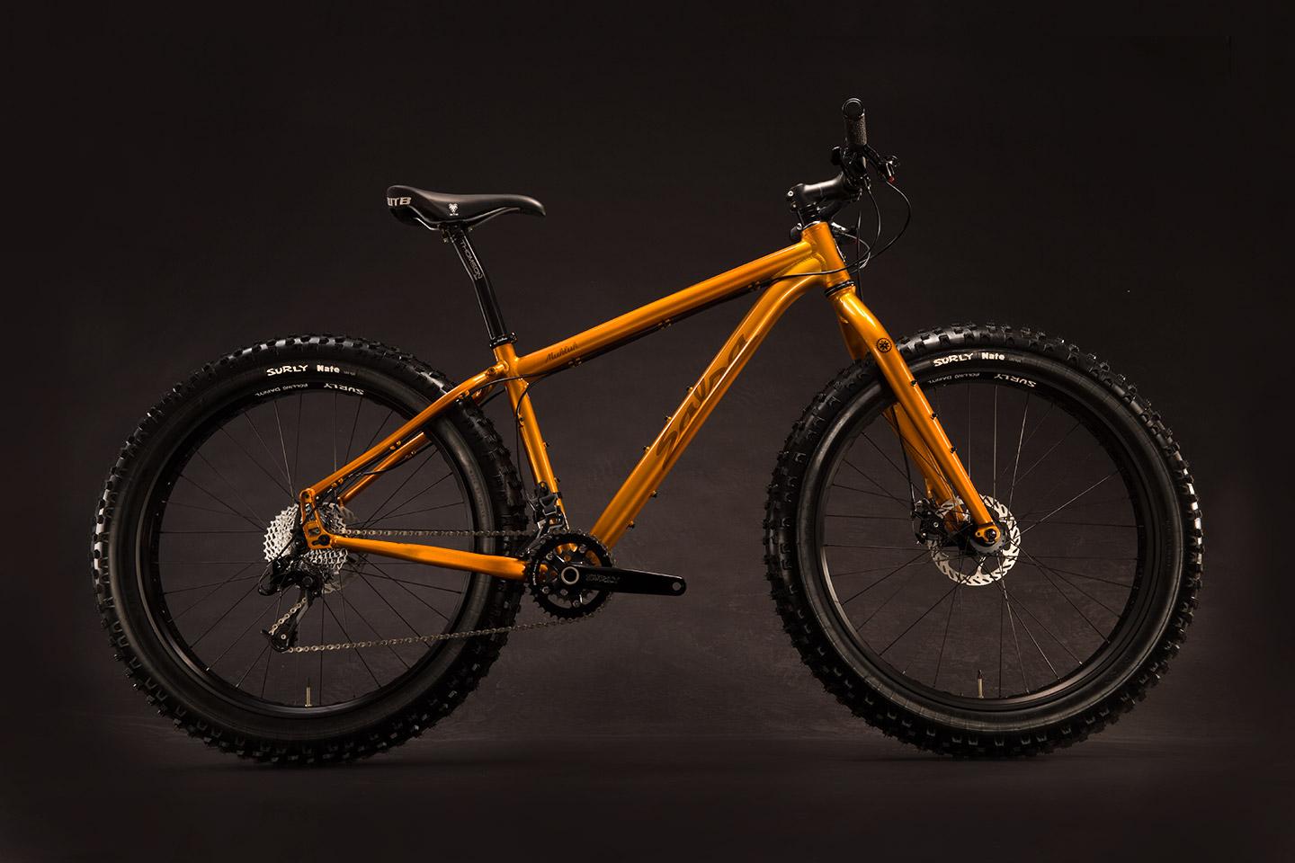 Salsa Mukluk: A vibrant orange mountain bike with wide tires, featuring a sturdy frame and a sleek design, set against a dark background. The bike is positioned sideways, highlighting its robust wheels and detailed components, including a gear system and handlebar.