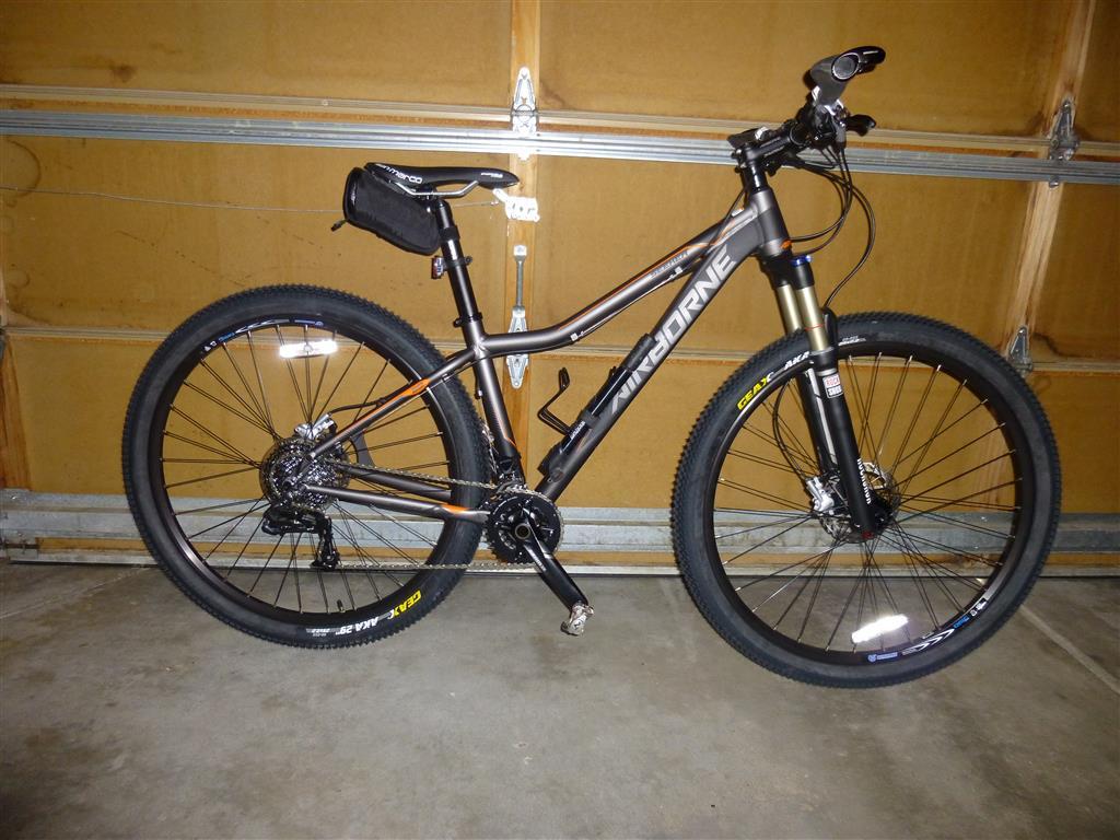 Airborne Seeker: A black and orange mountain bike with thick tires and a suspension fork, positioned upright in a garage with wooden walls and a concrete floor. A small bag is attached to the frame, and the bike features disc brakes and a gear shifter on the handlebars.