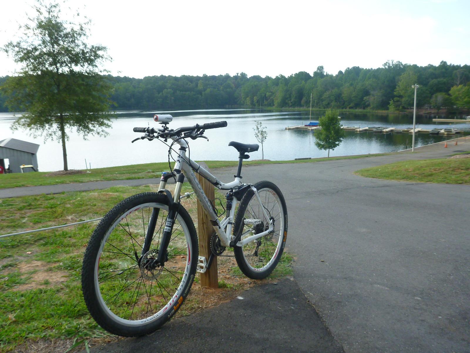 A mountain bike parked next to a lake with lush greenery in the background, featuring a small dock and a boat ramp. The scene evokes a serene outdoor environment, perfect for biking and enjoying nature. Salem Lake mountain bike trail.
