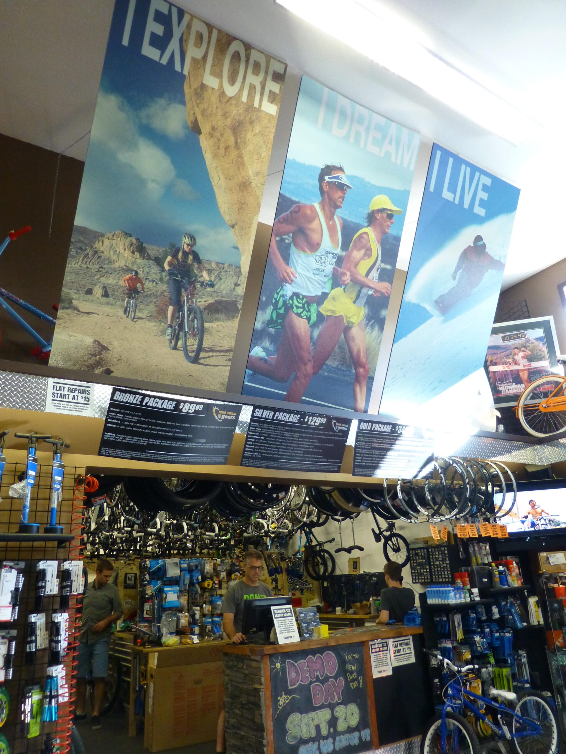 An interior view of a bike shop featuring promotional posters with the words "EXPLORE," "DREAM," and "LIVE," alongside images of people biking, running, and surfing. The shop is filled with bicycle accessories, tools, and equipment, with two staff members assisting customers at the checkout counter. A sign for "DEMO DAY" and pricing packages is visible on the counter.