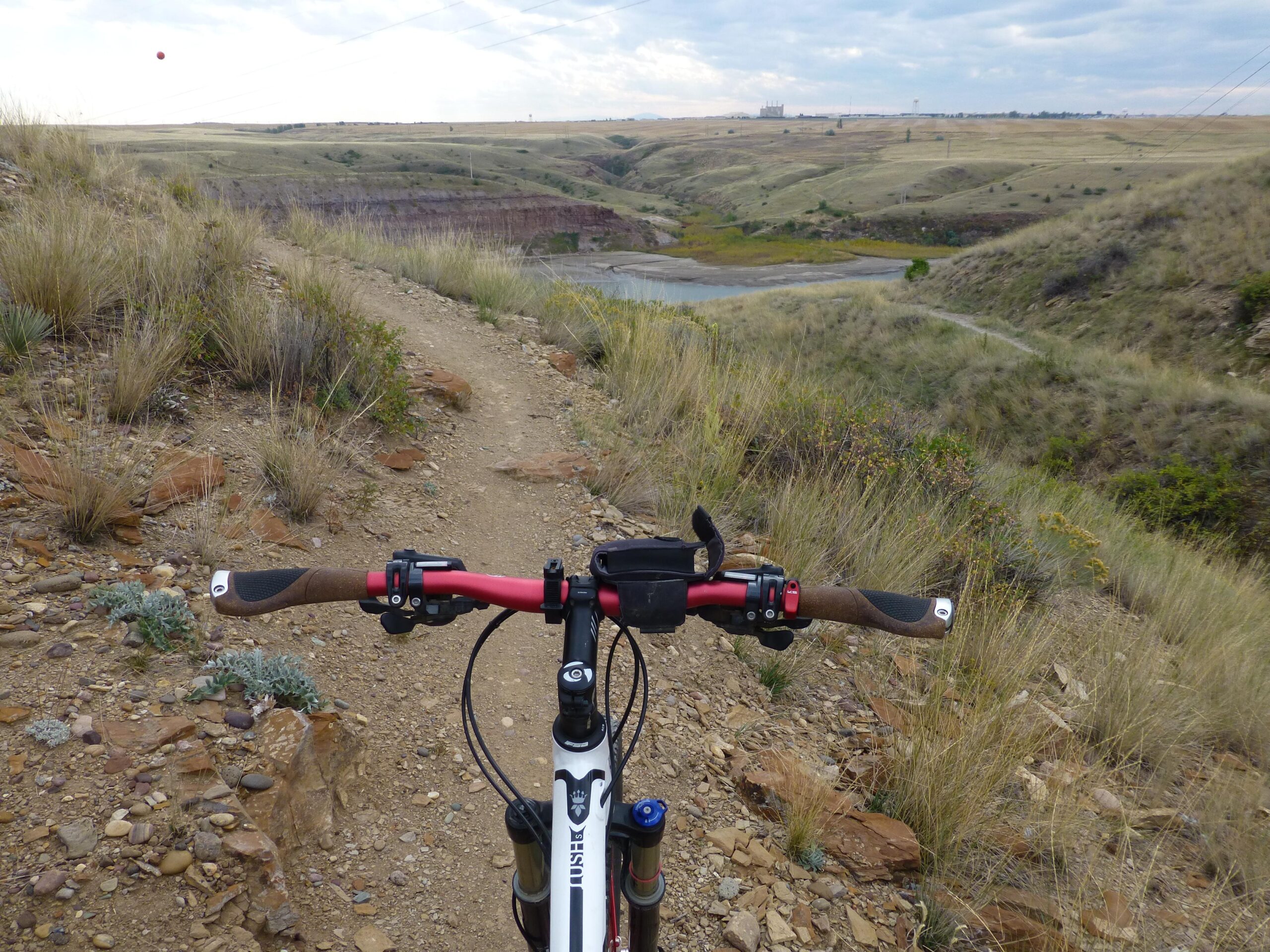 A close-up view of a mountain bike's handlebars, with a dirt trail winding through a hilly landscape in the background. The scene features dry grass, scattered rocks, and a distant view of a body of water and a cloudy sky above. New Video Game/Yeah Kid mountain bike trail.