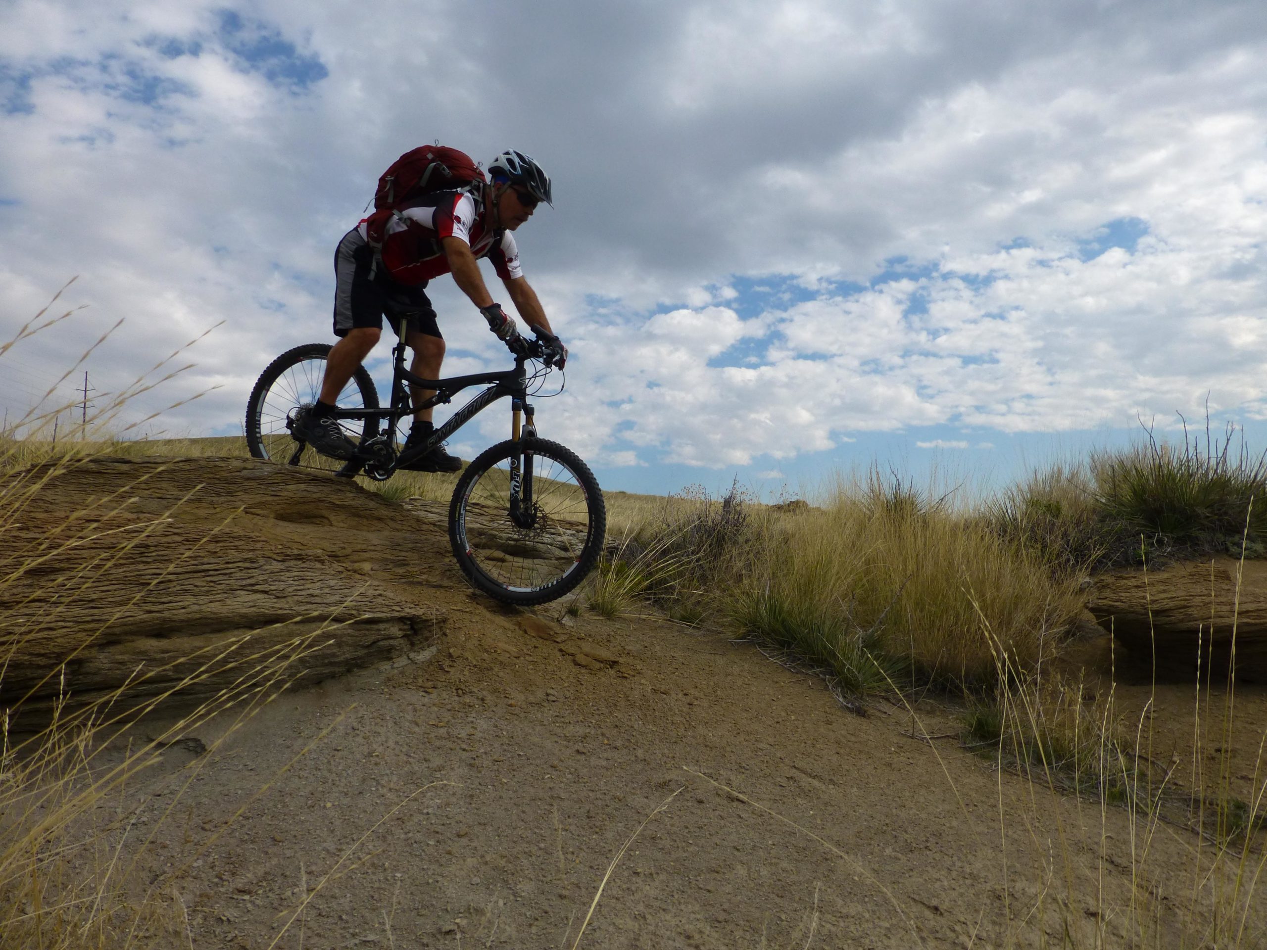 A mountain biker navigating a rocky terrain, leaning forward as they descend a slope. The rider is wearing a helmet and a backpack, surrounded by tall grass and a cloudy sky. New Video Game/Yeah Kid mountain bike trail.