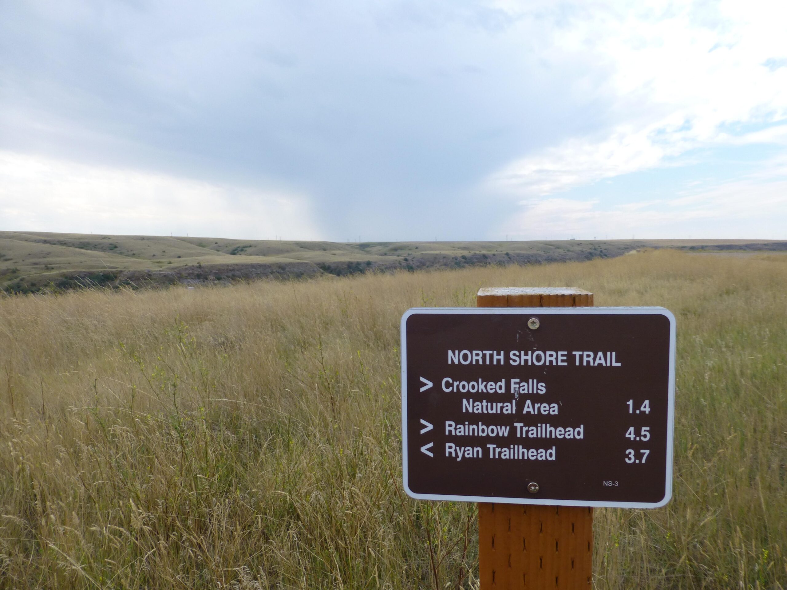 A signpost indicating the North Shore Trail, with directions to Crooked Falls (1.4 miles), Rainbow Trailhead (4.5 miles), and Ryan Trailhead (3.7 miles). The background features rolling hills under a cloudy sky, with tall grass in the foreground. New Video Game/Yeah Kid mountain bike trail.