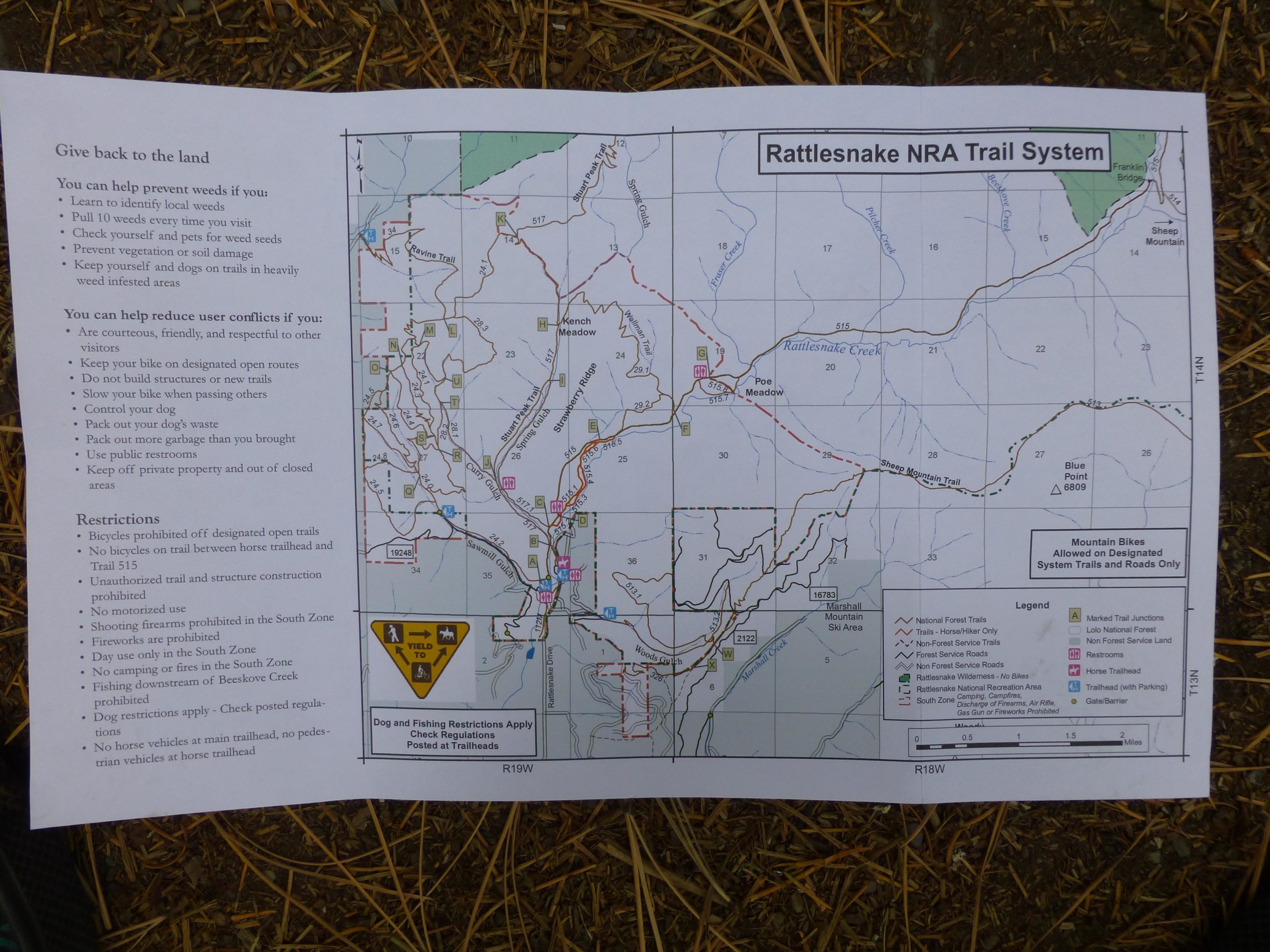 Map of the Rattlesnake National Recreation Area (NRA) Trail System, featuring trail routes, restrictions, and guidelines for visitors. The map includes notes on how to prevent weeds and reduce user conflicts, with a legend indicating various trail types, restrooms, and other facilities. The layout is surrounded by a natural setting, with pine needles and grass visible in the background. Rattlesnake mountain bike trail.