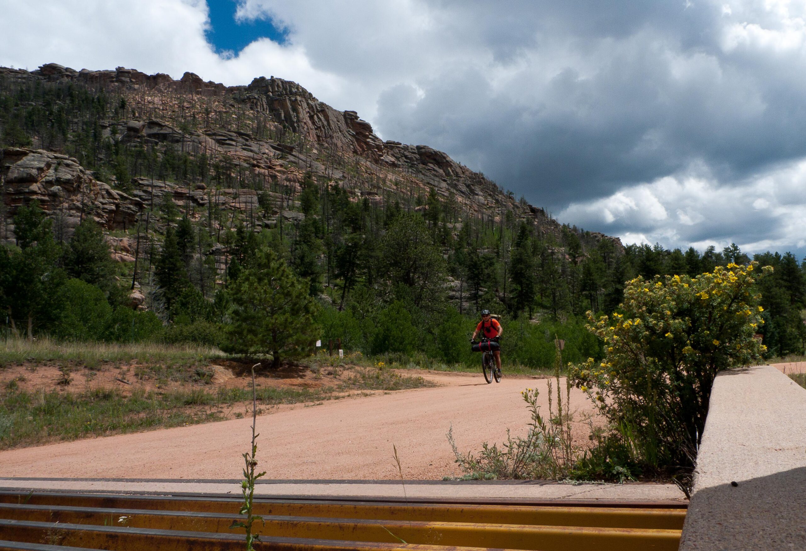 A cyclist riding on a dirt path surrounded by lush greenery and rocky hills under a partly cloudy sky. Colorado Trail mountain bike trail.