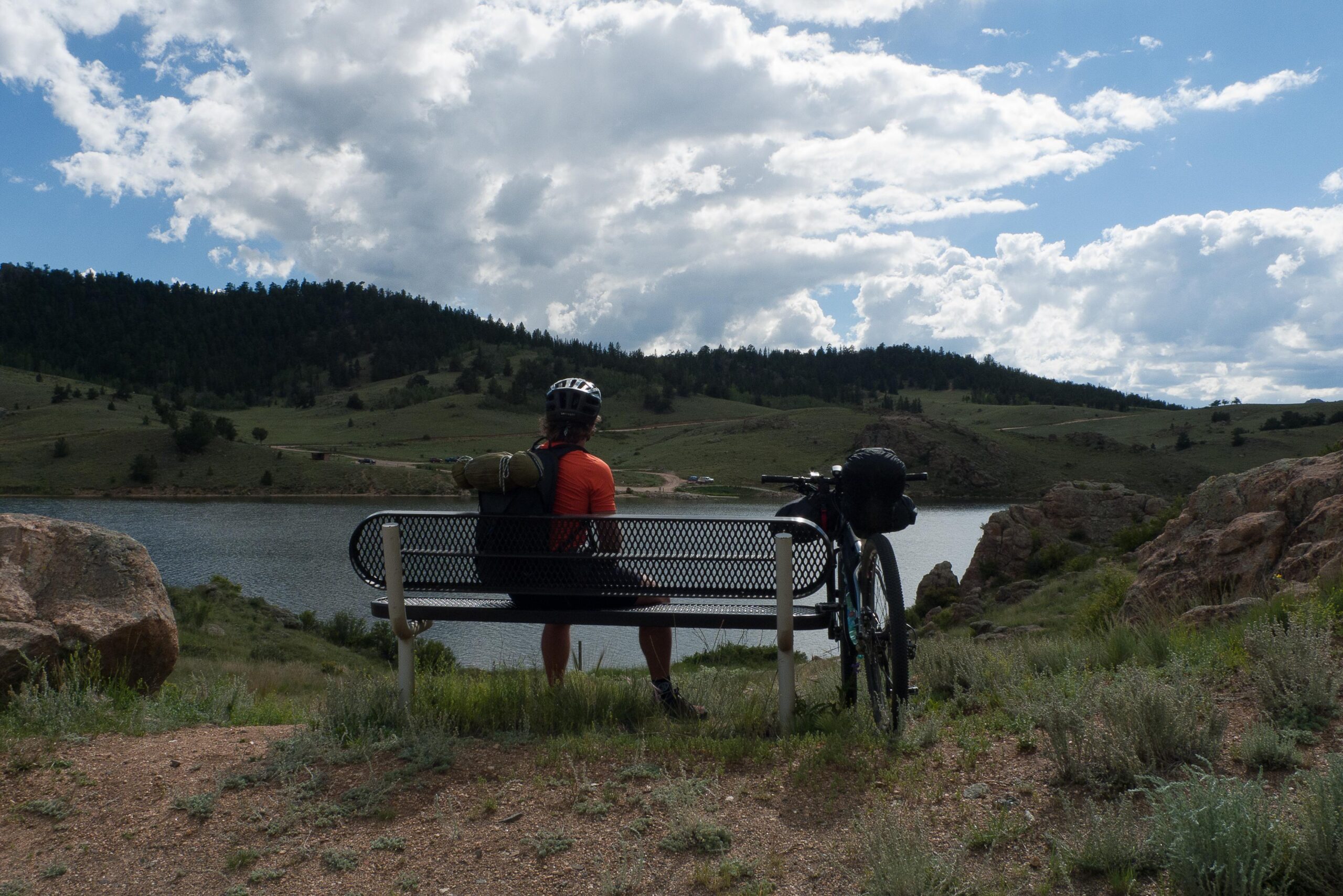 A cyclist sitting on a bench overlooking a serene lake surrounded by rolling hills and greenery. The cyclist, wearing a helmet and a backpack, gazes at the scenic landscape under a partly cloudy sky. A bicycle with gear is propped beside the bench. Colorado Trail mountain bike trail.