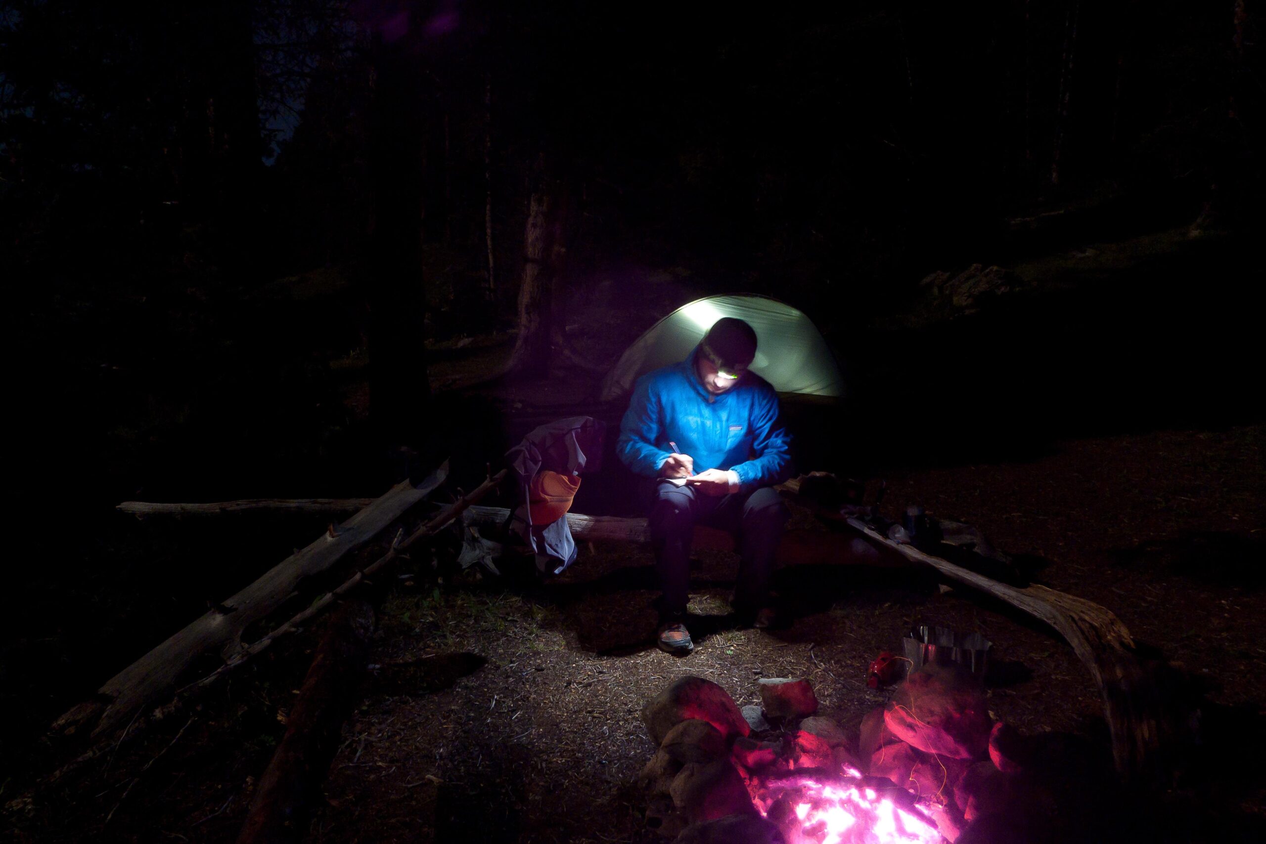 A person sitting by a campfire at night, wearing a blue jacket and reading or writing, illuminated by a headlamp. A green tent is set up in the background, and the glow of the fire casts warm light on the surroundings, highlighting logs and stones nearby. Colorado Trail mountain bike trail.