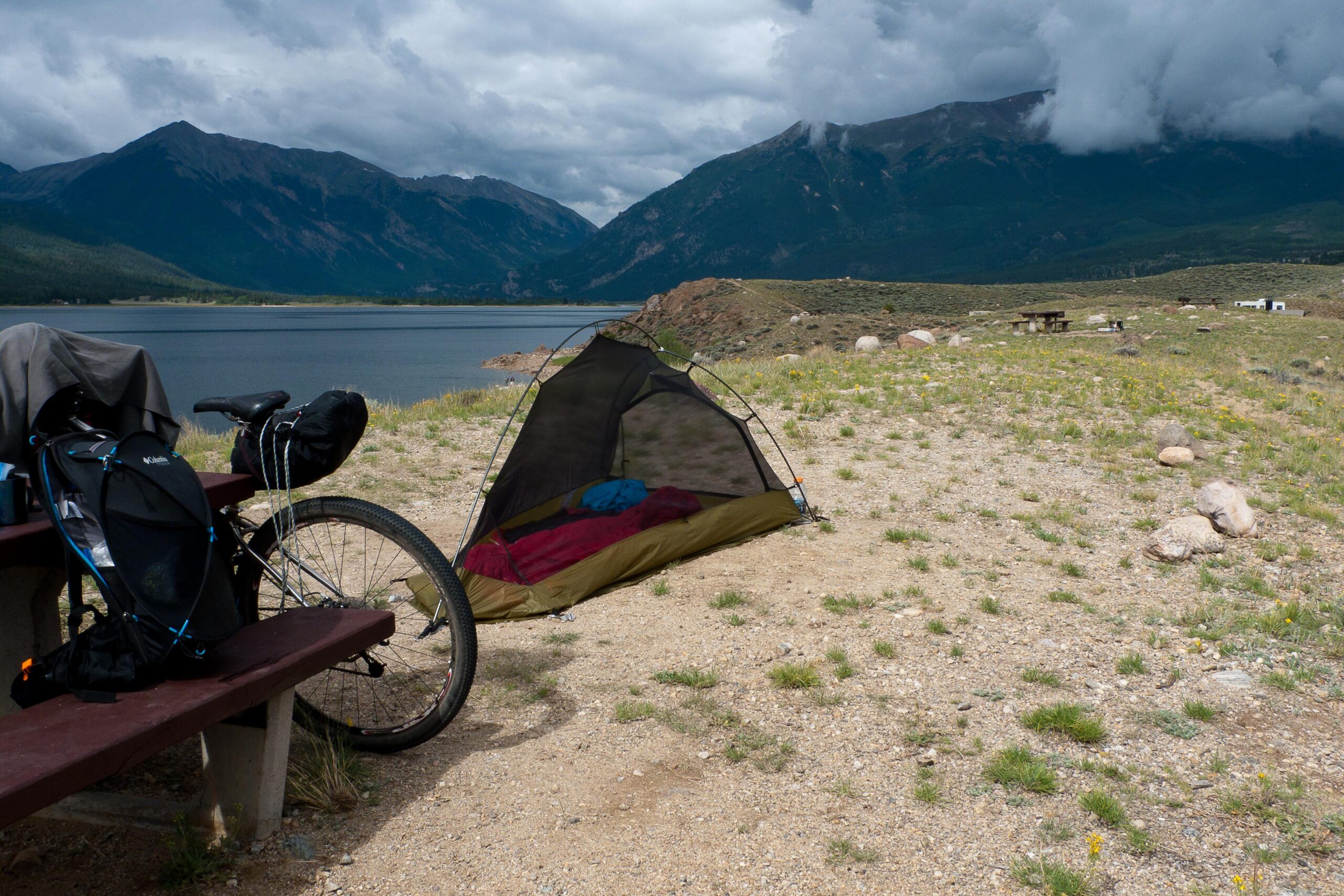 A camping scene by a lake featuring a bicycle, a backpack, and a small tent. Mountains rise in the background under a cloudy sky, with a picnic table and some rocky terrain visible nearby. The tent is pitched on sandy ground with a cozy sleeping bag inside. Colorado Trail mountain bike trail.