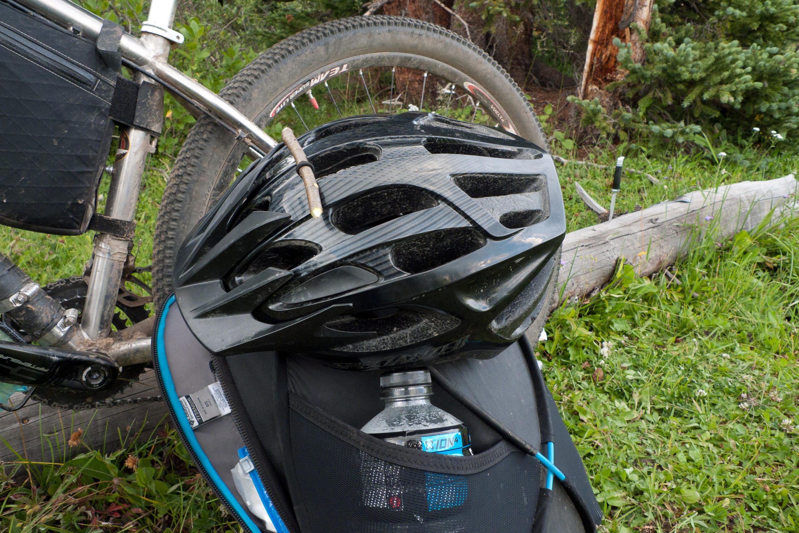 A black bicycle helmet resting on top of a backpack, with a water bottle partially visible from the bag. The bike's frame and wheel can be seen in the background, and the scene is set in a grassy area surrounded by trees. Colorado Trail mountain bike trail.