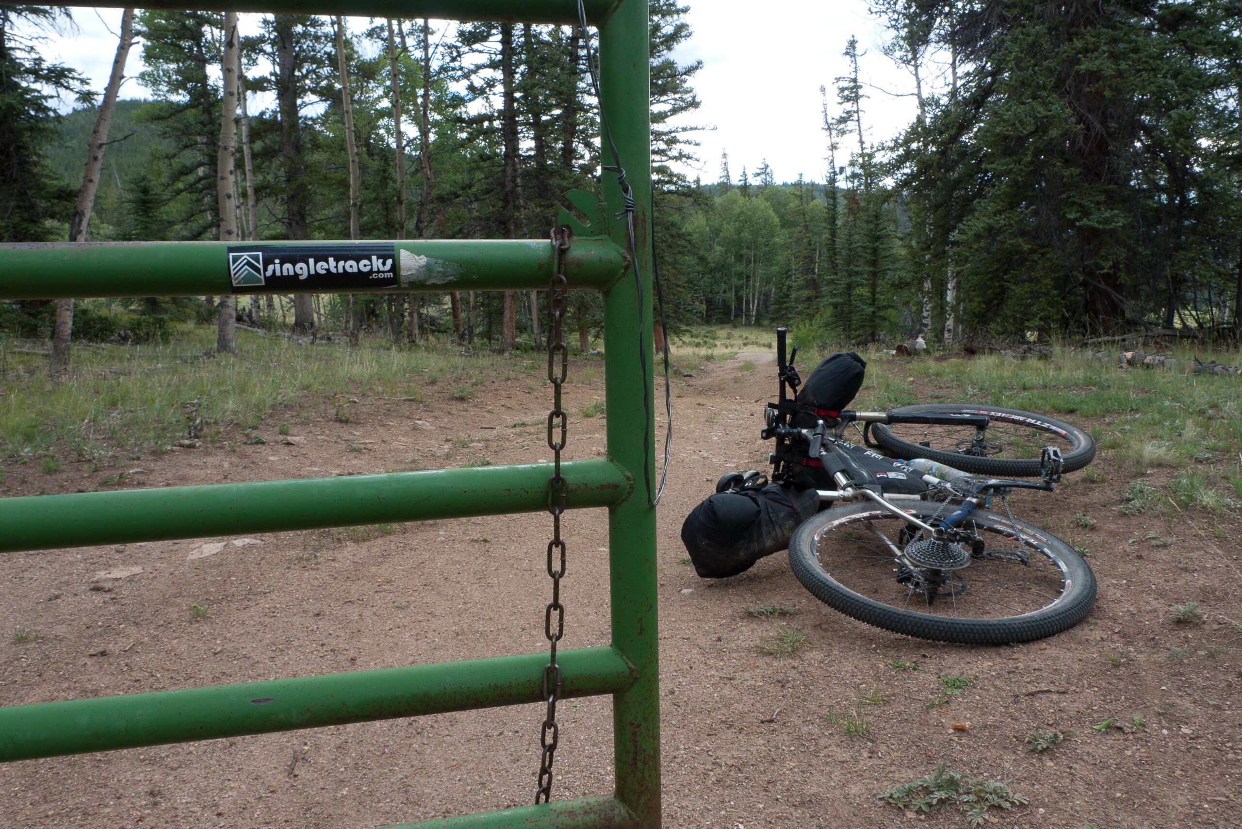 A green gate with a chain lies open, leading to a dirt path surrounded by trees. In the foreground, a mountain bike is on its side, with its wheels and gear visible. The setting is a natural area, suggesting a trail for biking or hiking. Colorado Trail mountain bike trail.