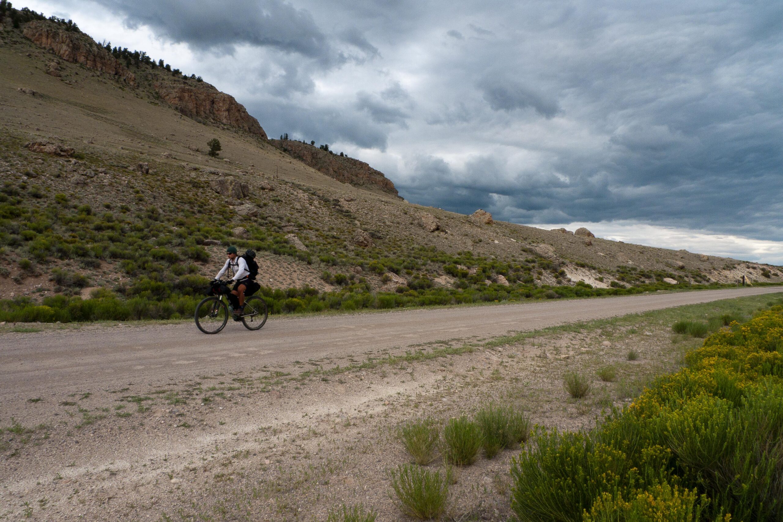 A cyclist rides along a gravel road surrounded by rolling hills and sparse vegetation under a cloudy sky. Colorado Trail mountain bike trail.