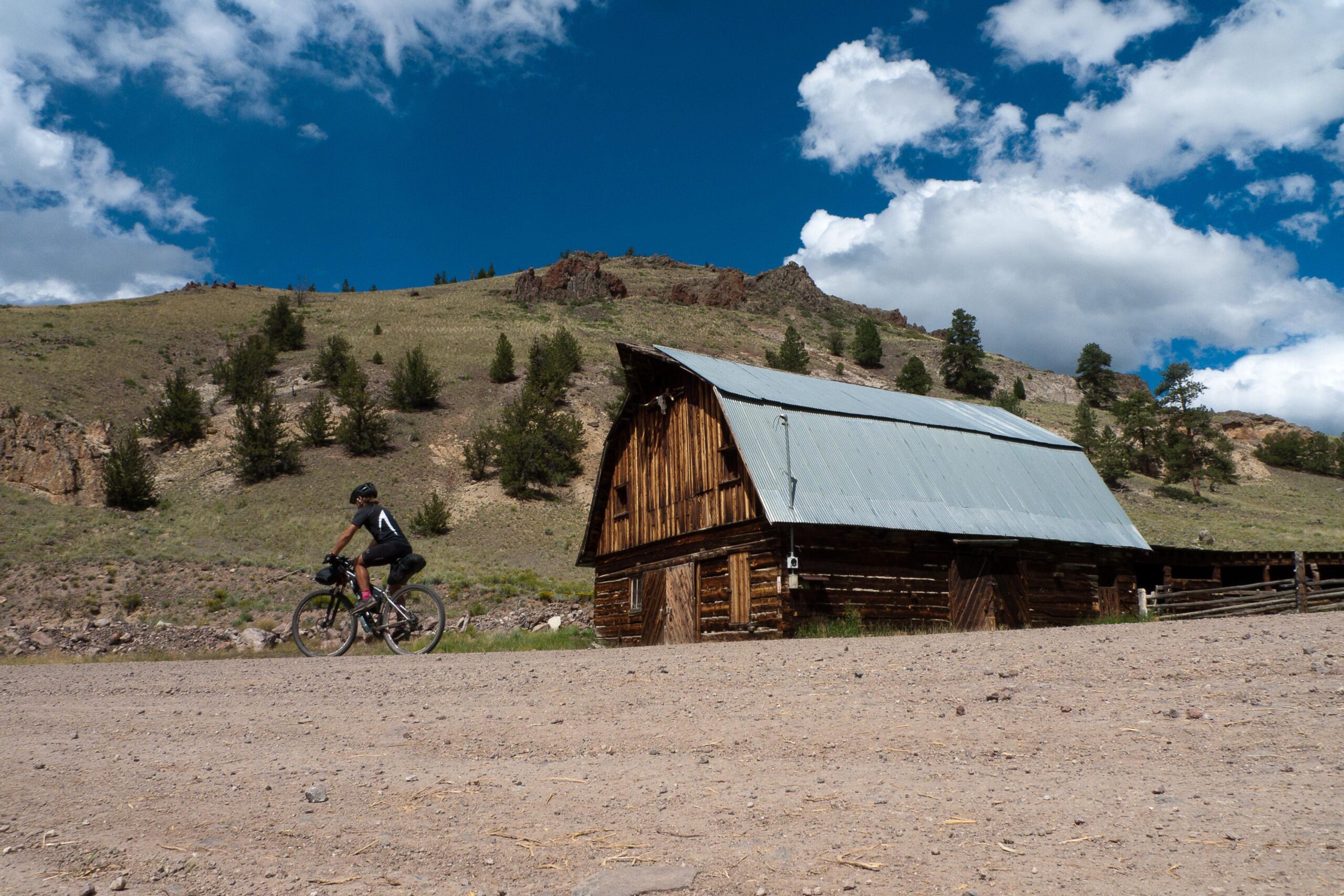 A cyclist rides along a dirt path beside a rustic wooden barn with a metal roof, set against a backdrop of rolling hills and a bright blue sky with scattered clouds. Pines dot the hillside, creating a peaceful rural landscape. Colorado Trail mountain bike trail.
