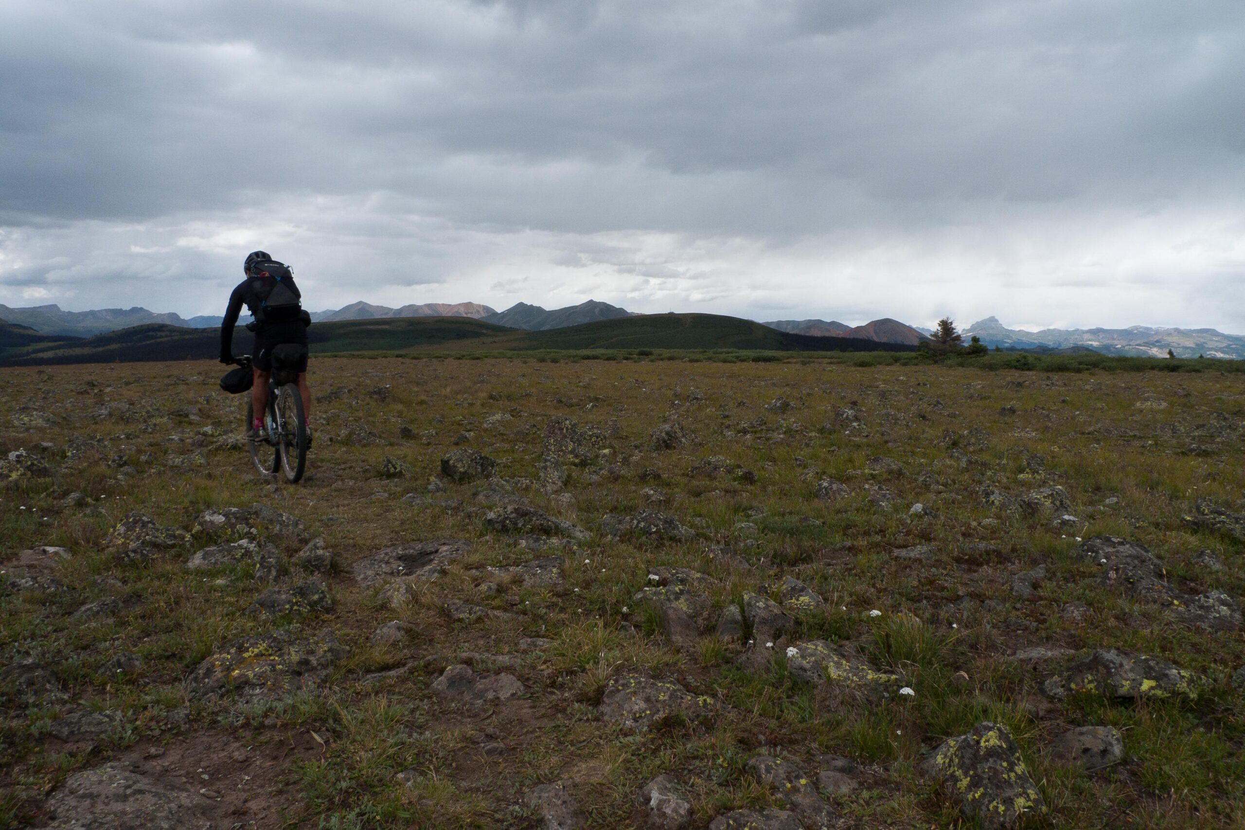 A cyclist riding a mountain bike through a rocky terrain under a cloudy sky, with rolling hills and distant mountains in the background. Colorado Trail mountain bike trail.