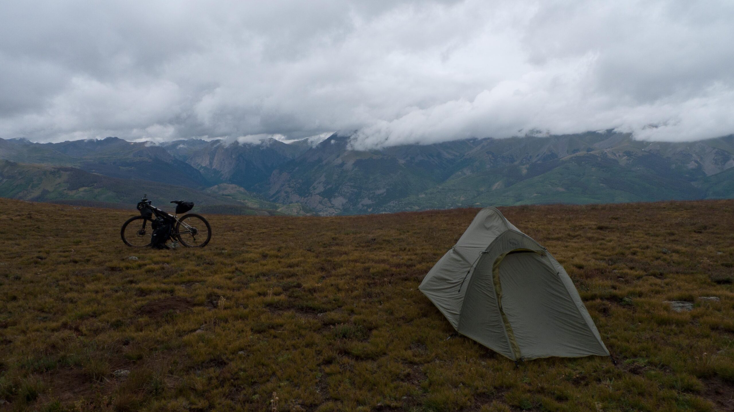 A bicycle parked on a grassy hillside with a green tent set up nearby, against a backdrop of cloudy mountains and an expansive sky. The scene captures a serene camping environment in a rugged outdoor landscape. Colorado Trail mountain bike trail.