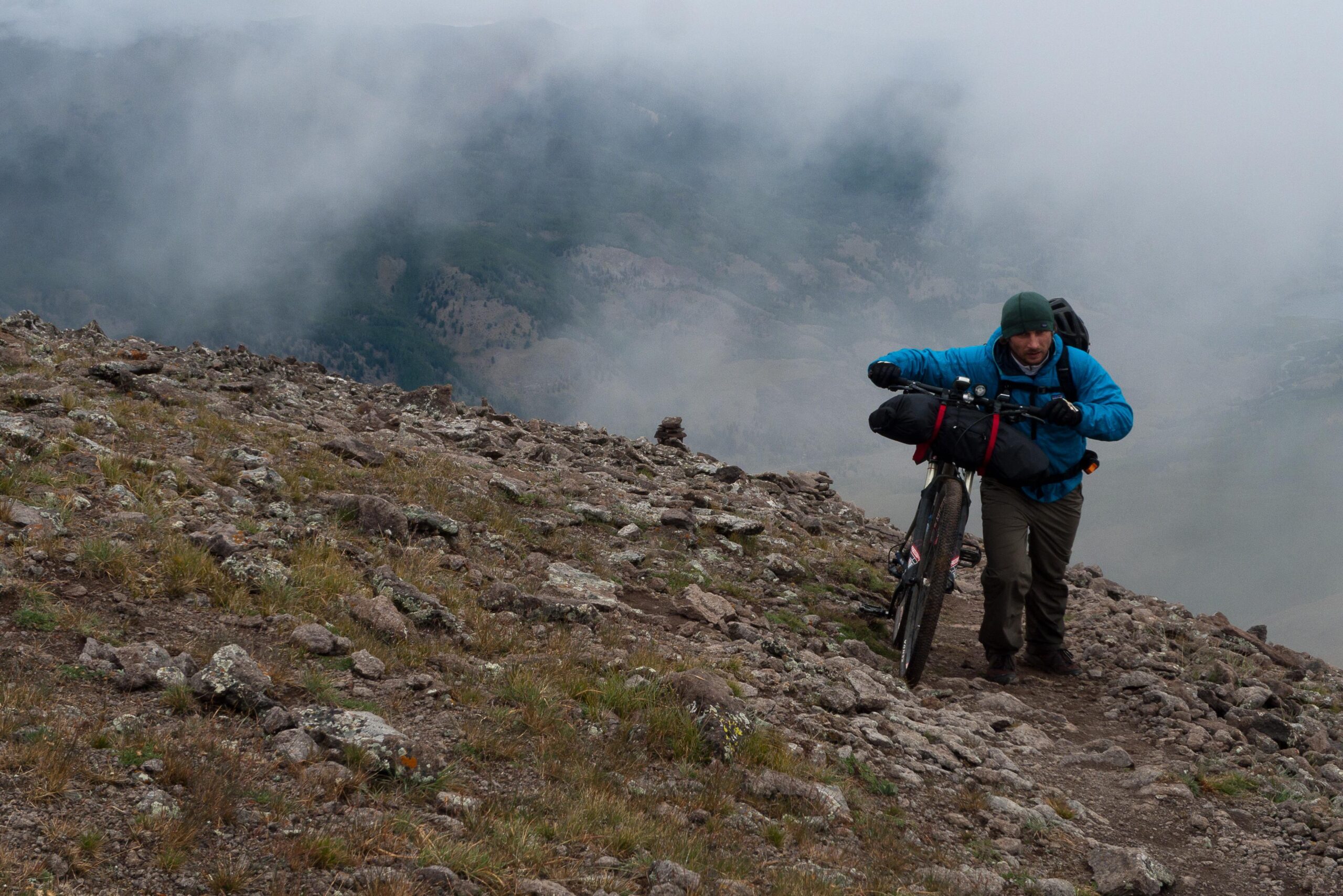 A person actively hiking up a rocky terrain while pushing a bicycle, surrounded by a misty and cloudy atmosphere. The individual is wearing a blue jacket and a green hat, with a backpack and gear attached to the bike. The landscape features rugged rocks and sparse vegetation, with a distant view of hills partially obscured by clouds. Colorado Trail mountain bike trail.