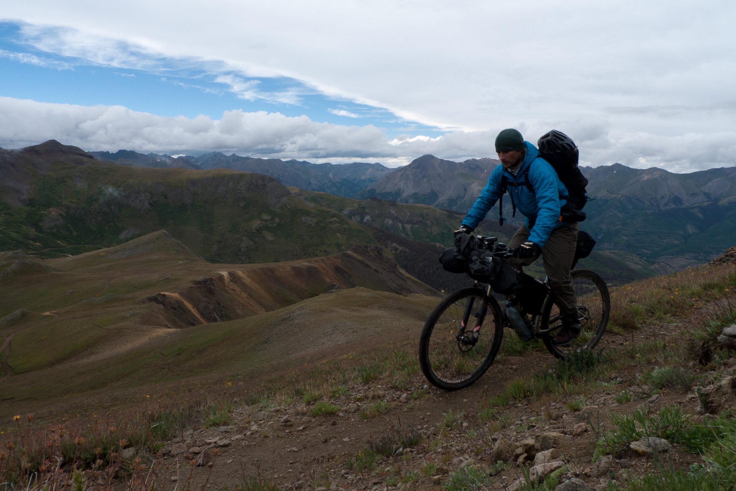 A cyclist navigating a rugged mountain trail, wearing a blue jacket and carrying a backpack. The landscape features rolling hills and distant peaks under a partly cloudy sky. Colorado Trail mountain bike trail.
