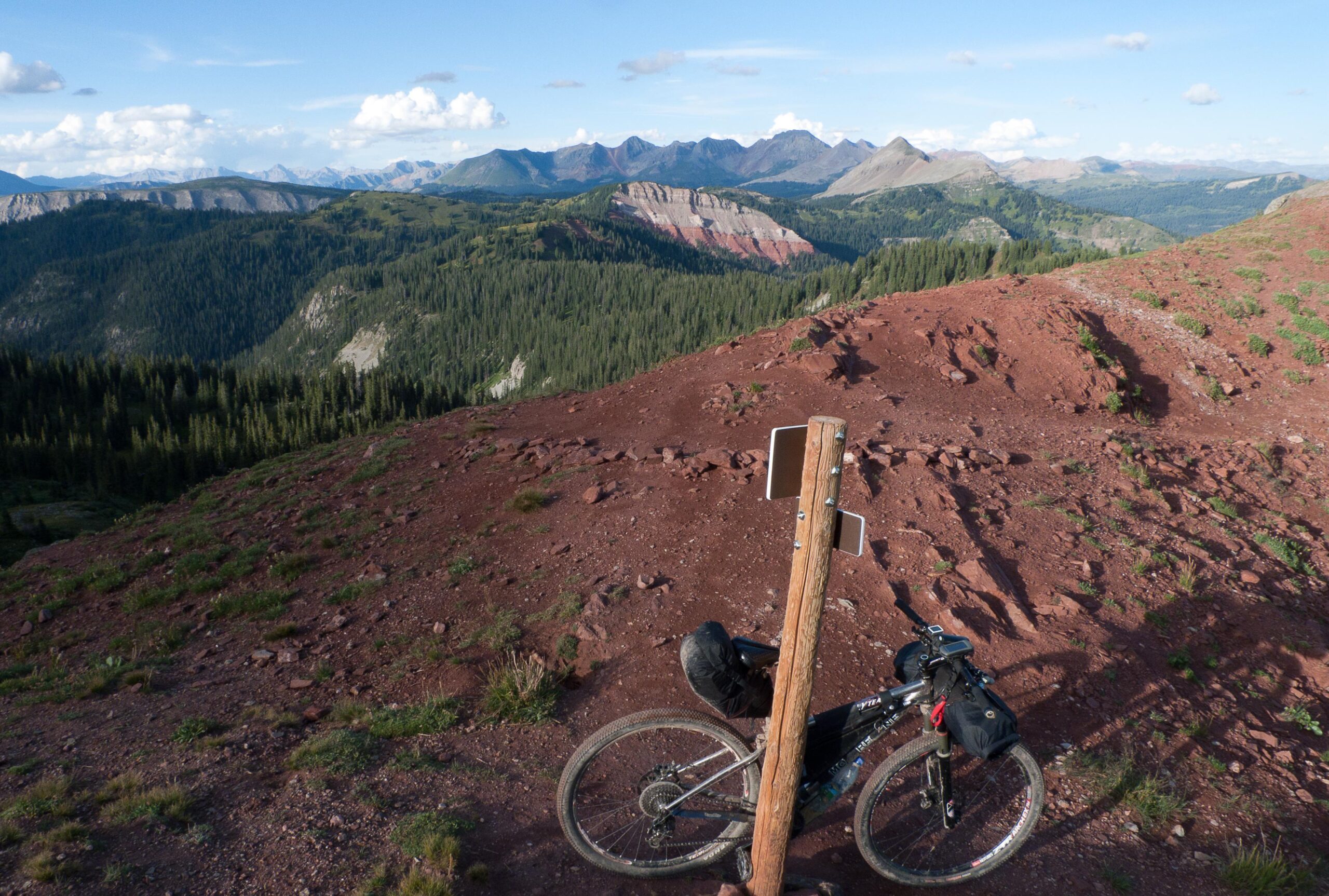 A scenic mountain landscape featuring a dirt path and a mountain bike parked next to a wooden post. The background showcases rolling hills, rugged peaks, and a clear blue sky with scattered clouds, surrounded by lush greenery and red soil. Colorado Trail mountain bike trail.